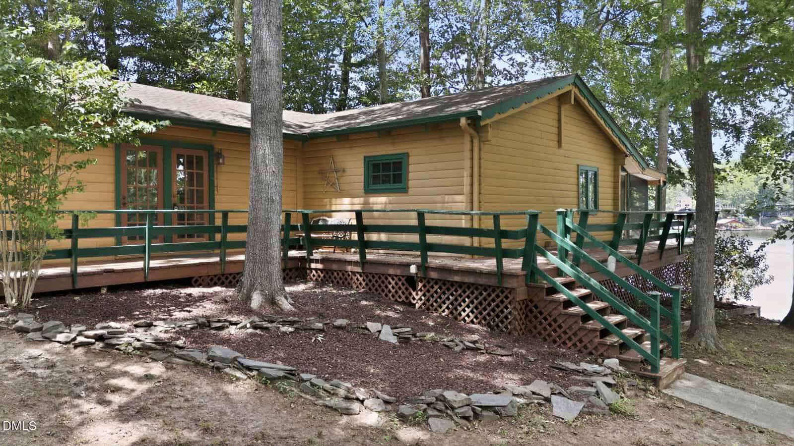 A rustic yellow log cabin with green trim features a ramp and steps leading to a wooden porch, surrounded by trees and a landscaped yard with rocks and mulch.