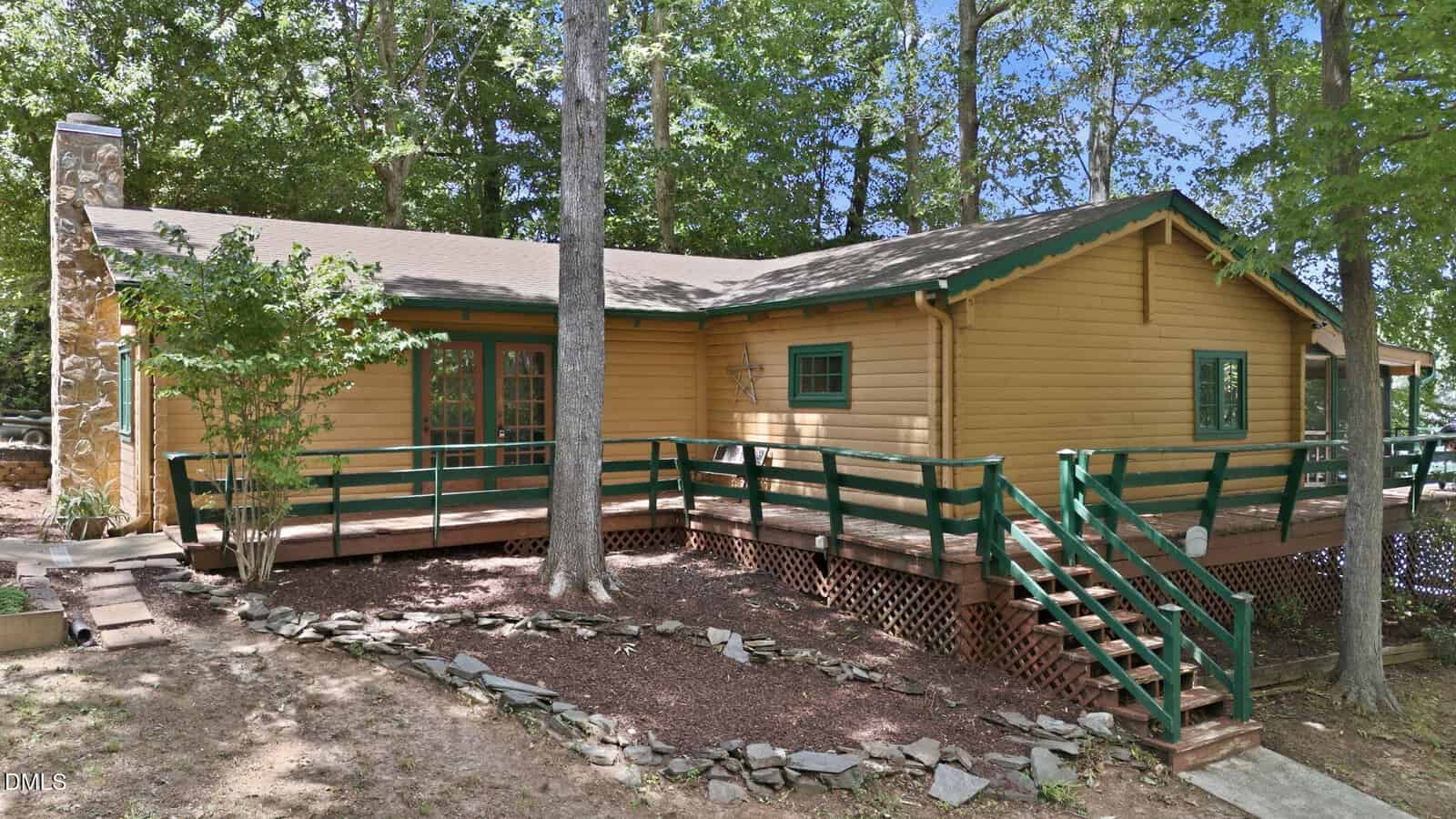 A yellow wooden cabin with green trim, a wraparound porch, green railings, and stairs. Surrounded by tall trees and a stone pathway, the cabin sits in a wooded area under a clear sky.