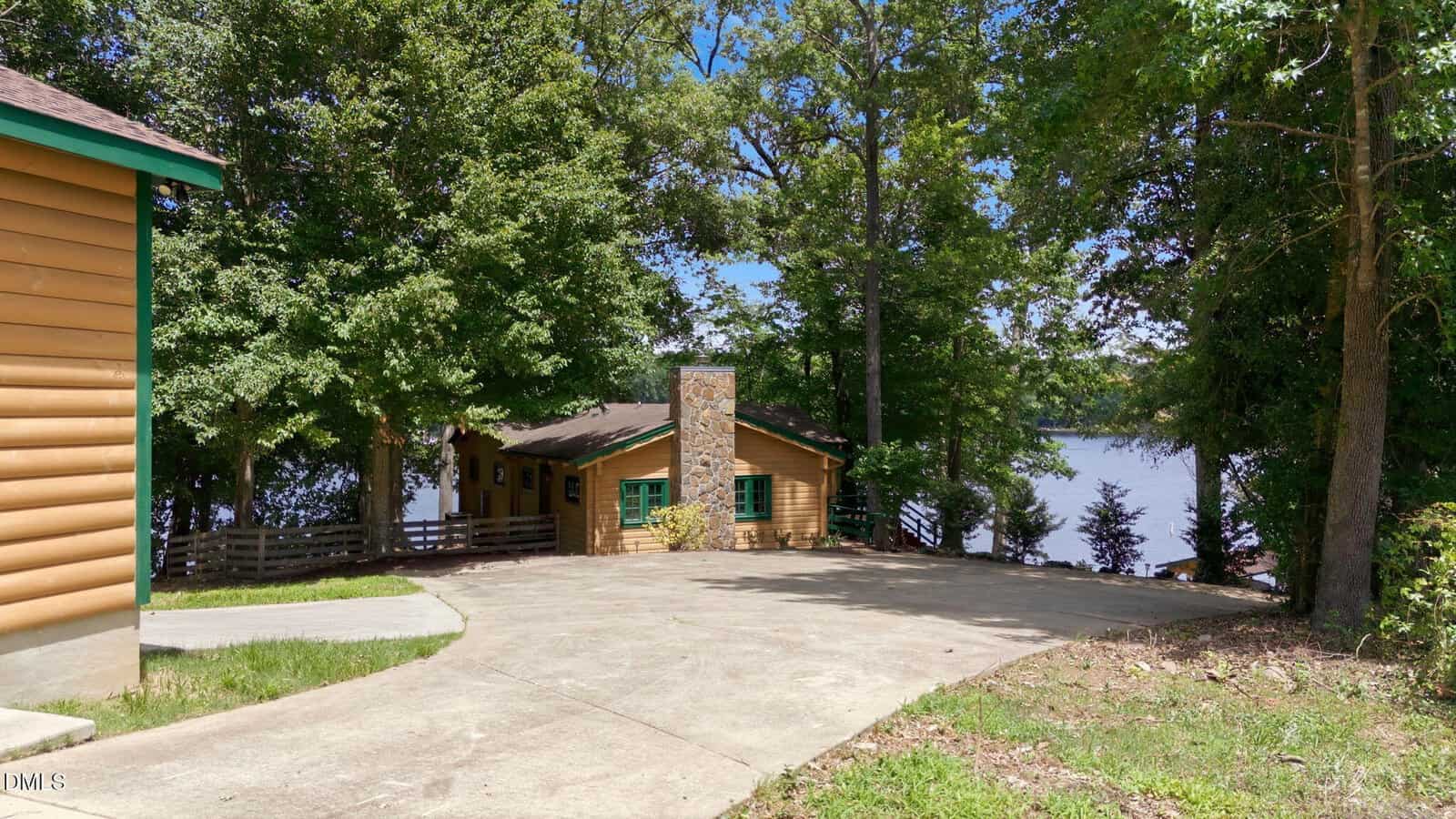 A wooden cabin with a stone chimney sits by a lake, surrounded by trees. A concrete driveway leads down to the cabin and the water, with sunlight filtering through the foliage.