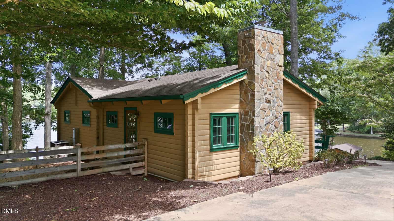 A small wooden cabin at 407 Sagamore Drive in Louisburg features tan siding, green-trimmed windows, and a stone chimney. Nestled among trees near water, it boasts a gravel driveway and wooden fence out front.