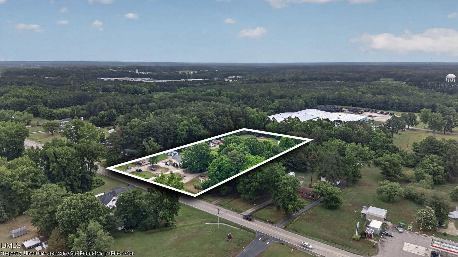 Aerial view of a tree-covered property outlined in white within a semi-rural area, surrounded by other houses, roads, open fields, and wooded land under a partly cloudy sky.