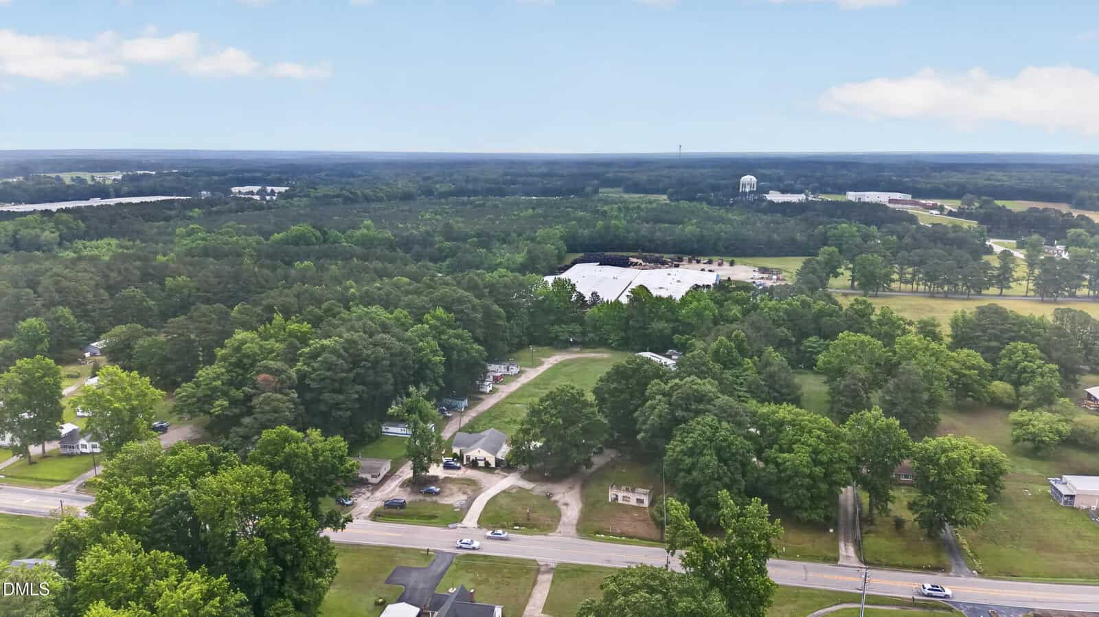Aerial view of a rural area with scattered houses, dense green trees, a large industrial building in the background, and a water tower visible on the horizon under a partly cloudy sky.