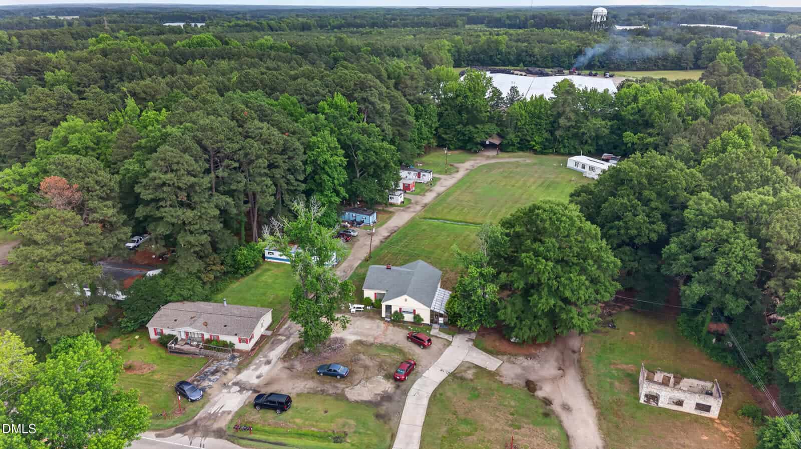 Aerial view of a rural area with several houses surrounded by trees, a grassy field, and a curving driveway. In the distance, smoke rises near a large white building and a water tower.