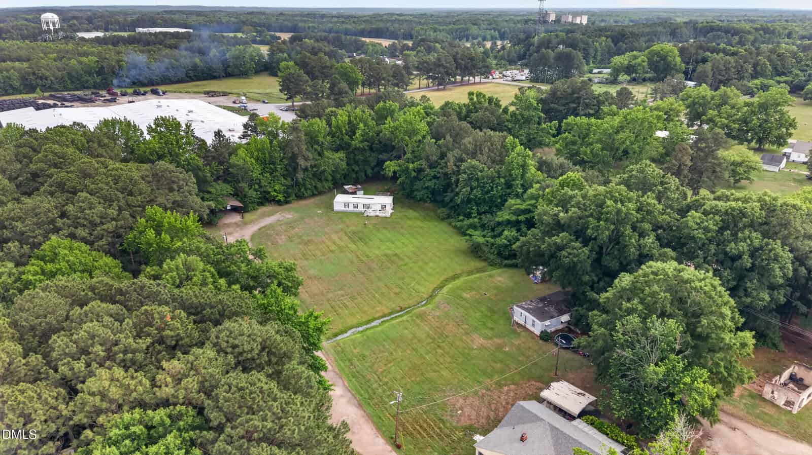 Aerial view of a rural area with a white house in the center, surrounded by trees and open grassy fields. Several other buildings are visible nearby, along with a large warehouse in the background.