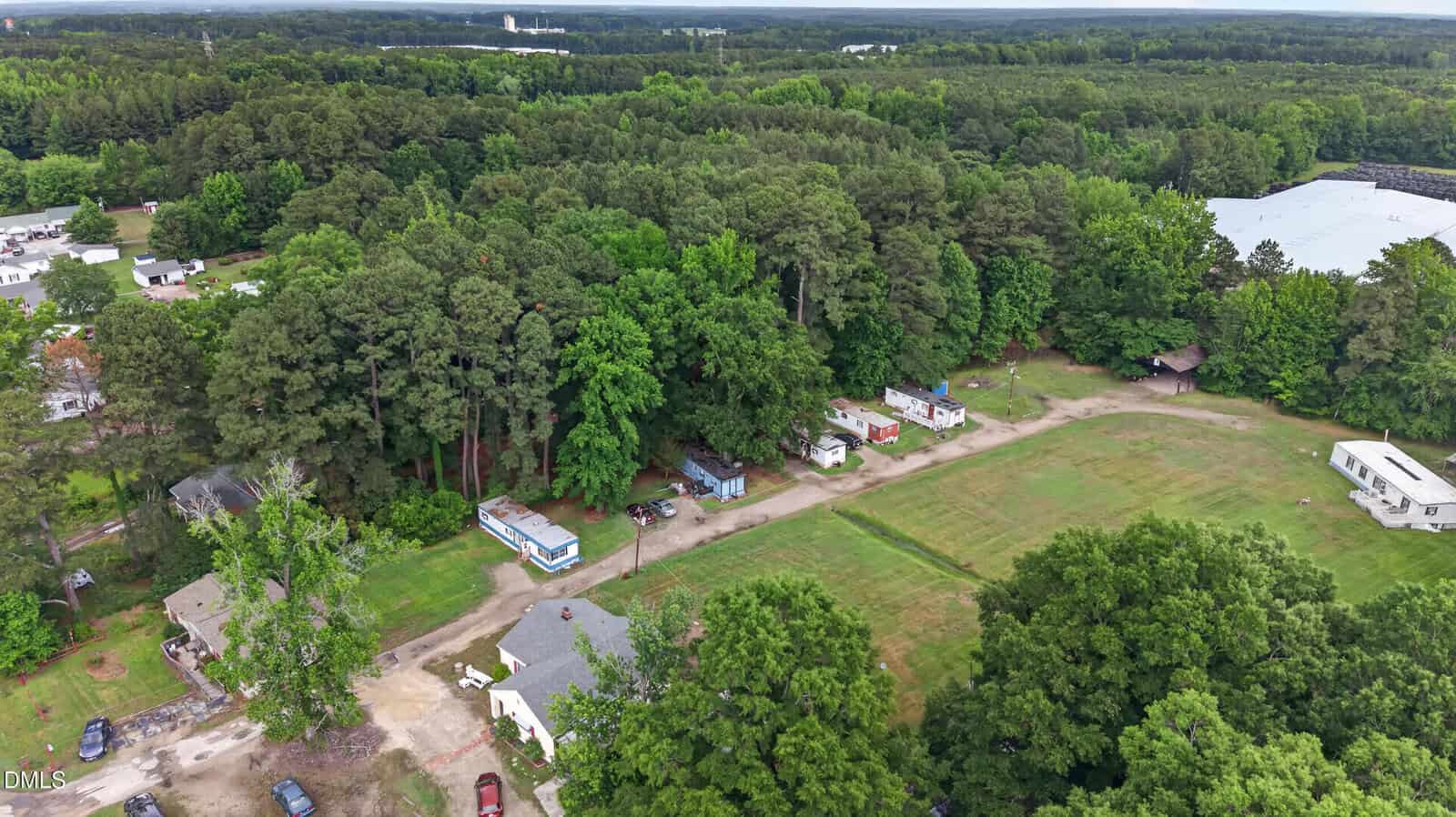 Aerial view of a rural area with scattered mobile homes, dirt roads, dense green trees, a large white building, and grassy open spaces under a partly cloudy sky.