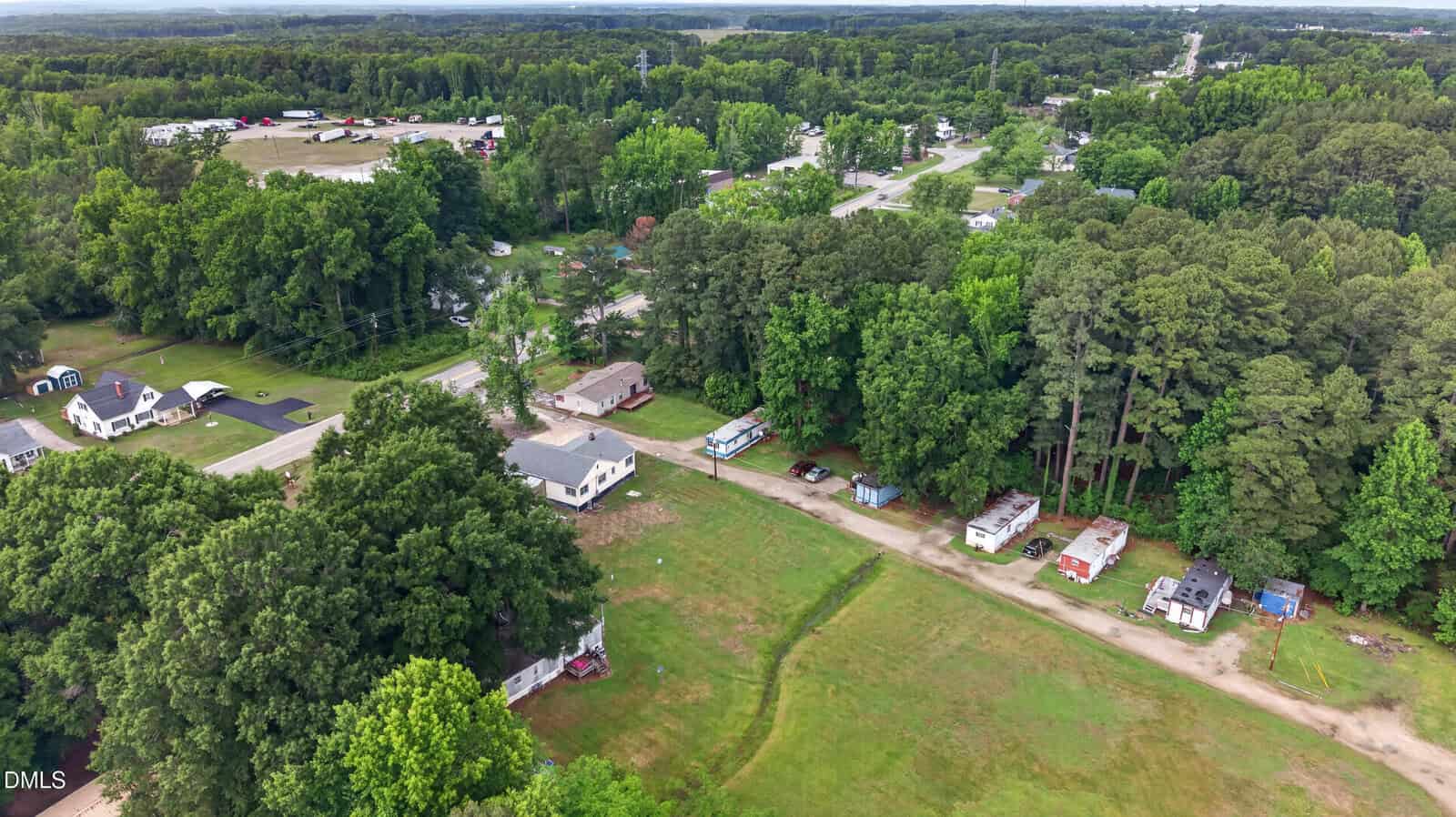 Aerial view of a rural area with scattered houses, trailers among trees, a main road, grassy fields, and dense forest in the background.