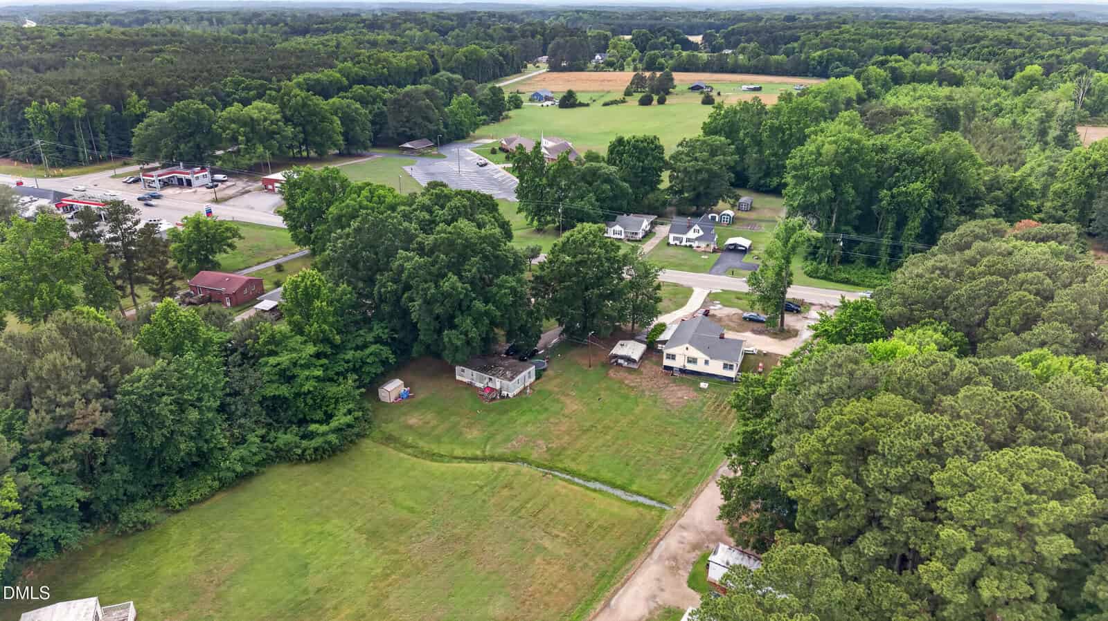 Aerial view of a rural area with scattered houses, mobile homes, grassy yards, dense trees, and a nearby commercial area with parking lot and roads. Farmland and forest surround the community.