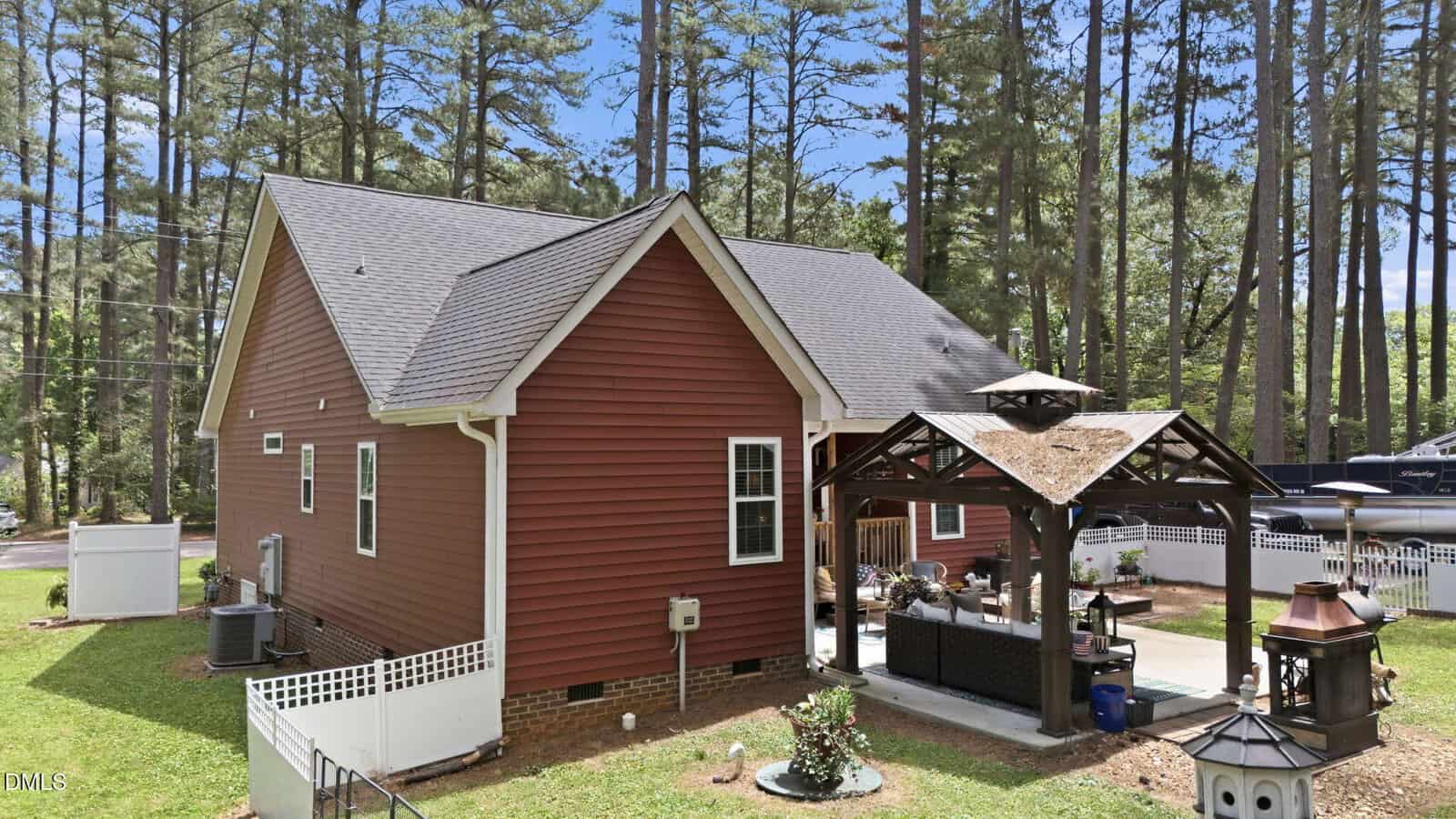 A red house with white trim is surrounded by tall pine trees. The backyard features a covered patio area with outdoor furniture, a fire pit, and a small garden, all set on a well-kept lawn.