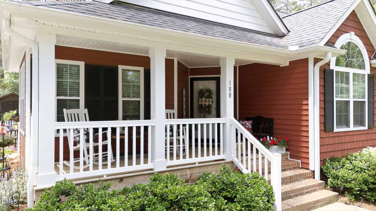 A charming red house with white trim at 188 Pine Cone Drive in Oxford features a front porch with rocking chairs, potted flowers, a welcome mat, a wreath on the door, and steps leading up to the entrance.
