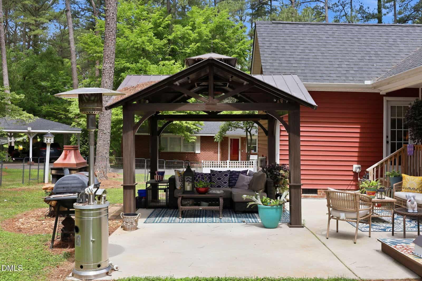 A backyard patio features a wooden gazebo with outdoor seating and cushions, a heater, grill, and chairs on a concrete surface. The house has red siding, surrounded by trees and greenery.