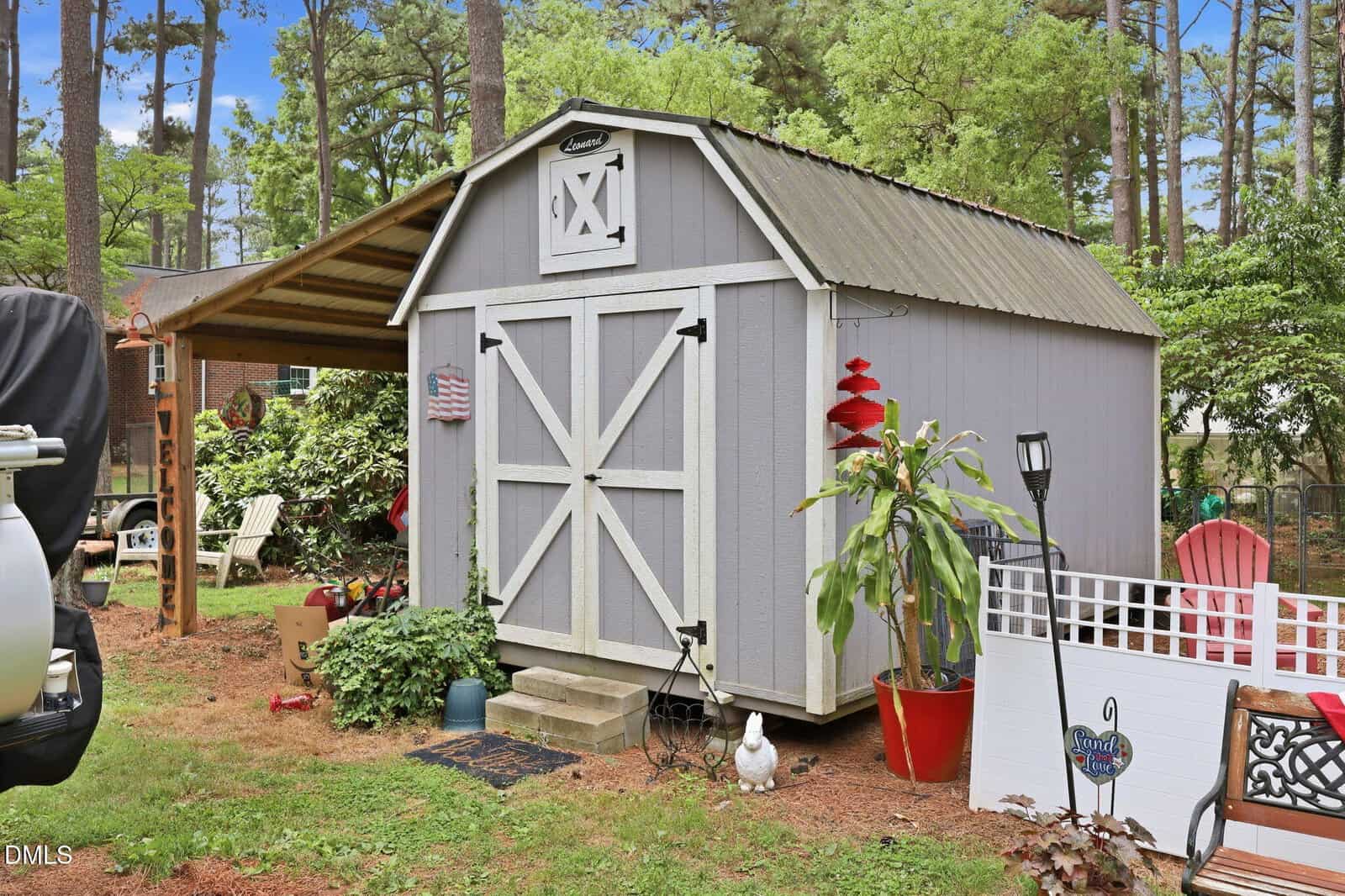 A gray barn-style shed with white trim sits in a backyard garden, surrounded by potted plants, a small staircase, lawn chairs, a white fence, and trees in the background.