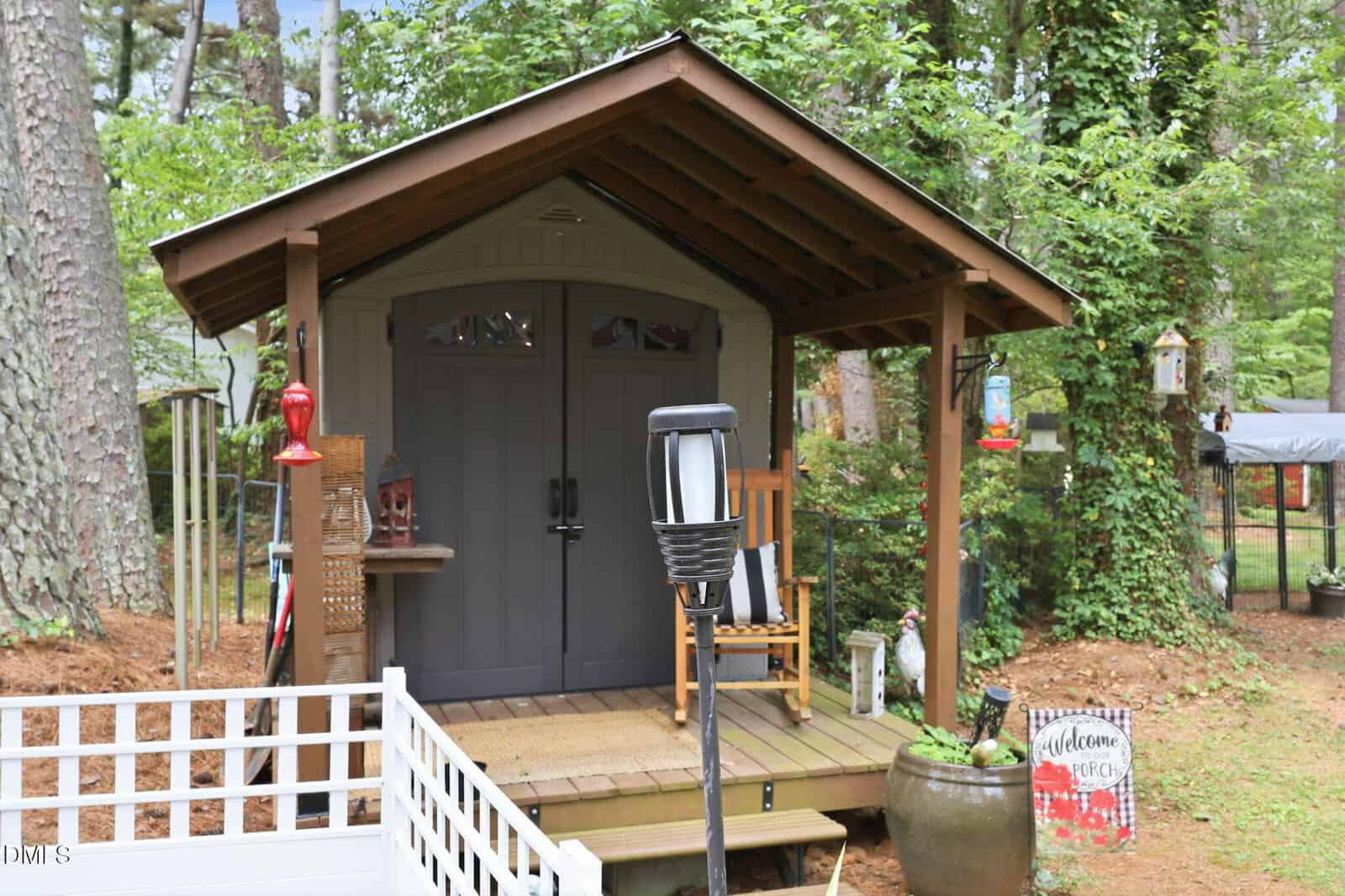 A small shed with double doors sits on a wooden porch, featuring a rocking chair and lamp post surrounded by trees, plants, and garden decor, including a “Welcome” sign and birdhouses.