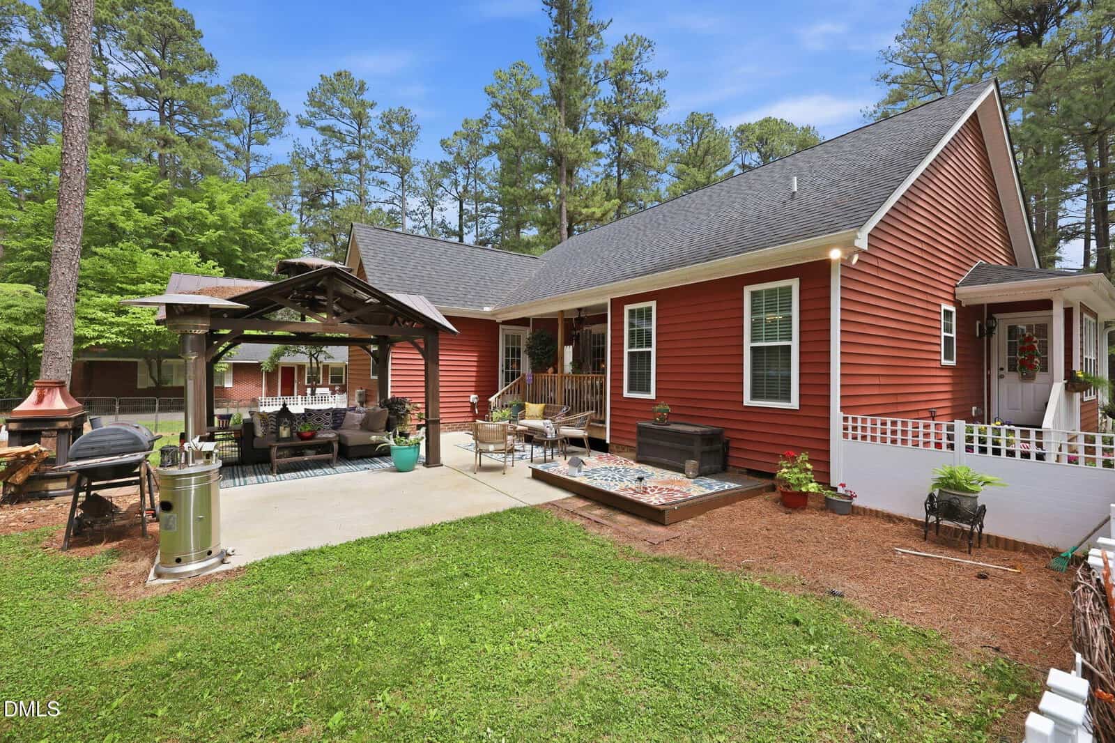 A red house with white trim sits amid tall trees. The backyard features a covered patio with seating, a fire pit, a small garden bed, and a grassy lawn under a blue sky.