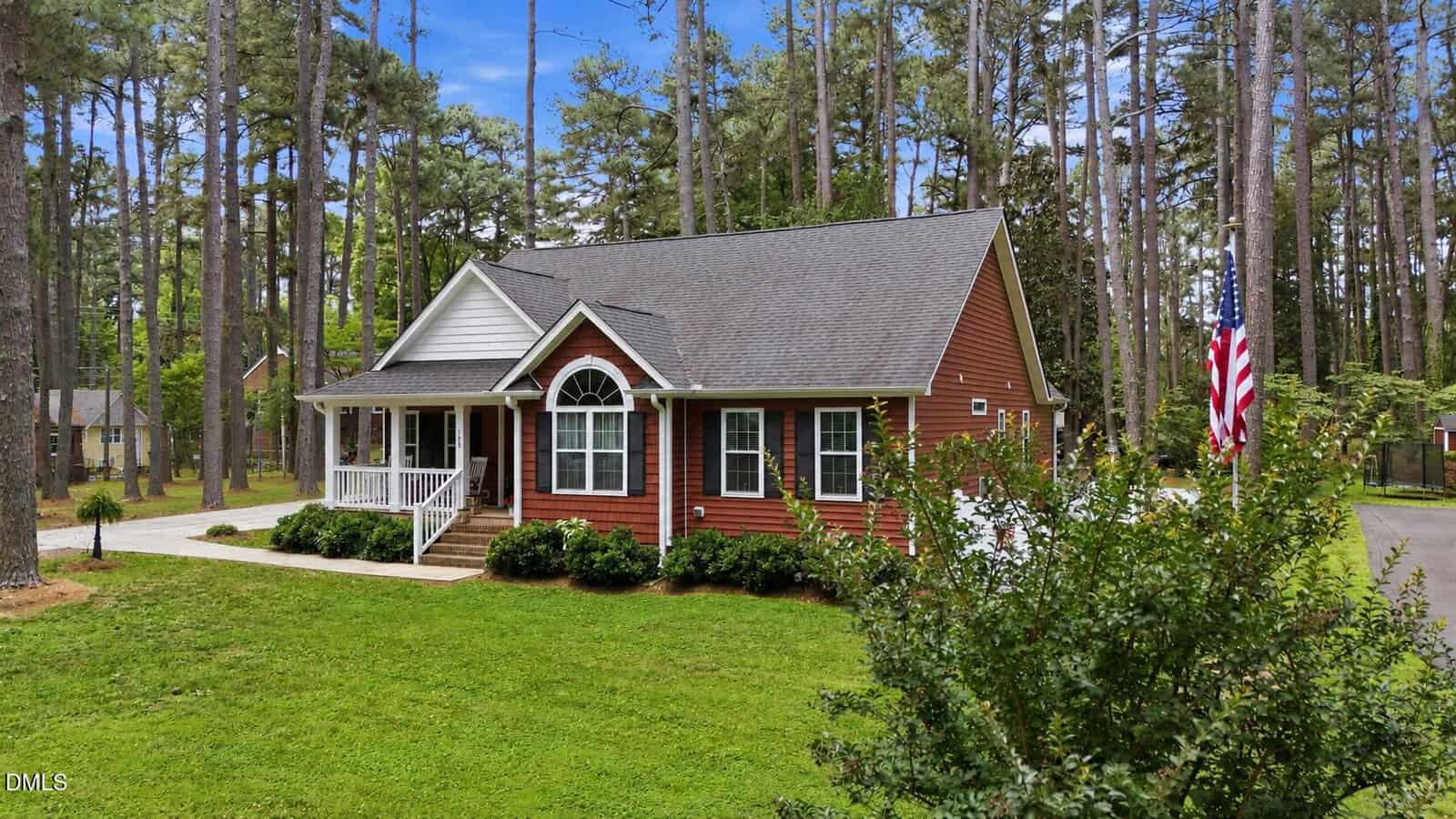 A single-story red house with white trim and a front porch sits on a grassy lawn at 188 Pine Cone Drive, Oxford, surrounded by tall pine trees. An American flag is displayed on the right, and the sky is blue with scattered clouds.