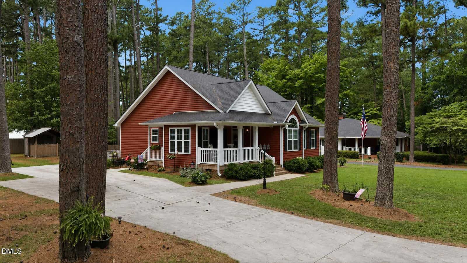 A charming red house with white trim and a covered front porch sits among tall pine trees, featuring a well-kept lawn and a long concrete driveway under a bright blue sky.