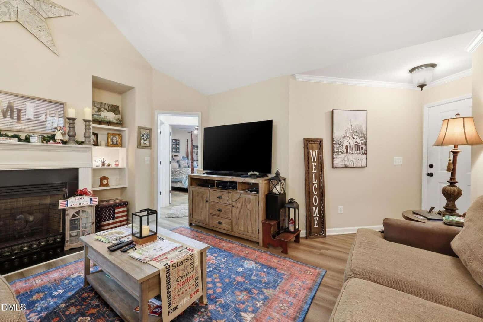 Cozy living room with tan walls, a fireplace, large TV on a wooden stand, brown sofa, coffee table, blue-red rug, and a “Welcome” sign on the wall. Room is warmly lit, with decor on shelves and a lamp by the sofa.