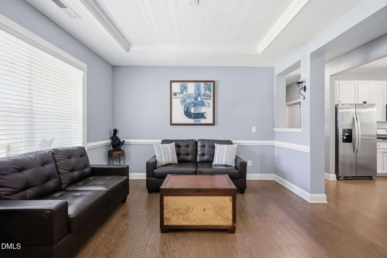 A modern living room with light blue walls, two dark brown leather sofas, a wooden coffee table, a small sculpture, and a framed abstract artwork. The space has large windows and an open view into a kitchen.