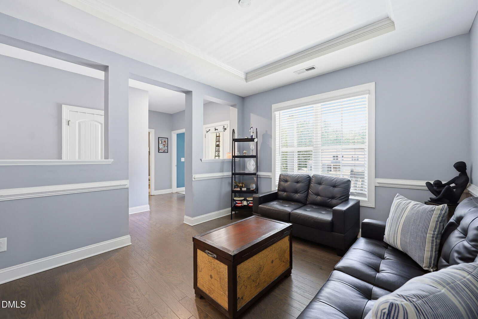 A bright living room with light blue walls, dark wood floors, two black leather sofas, a wooden chest coffee table, a shelf with storage bins, and a large window with white blinds letting in natural light.