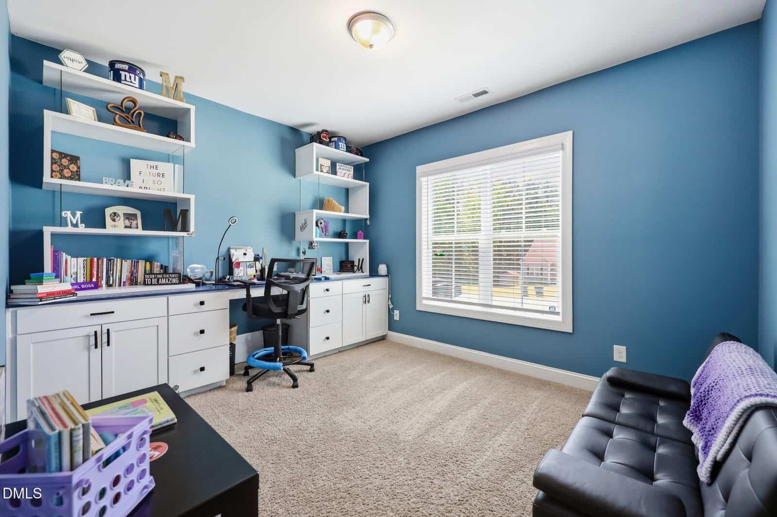 A home office with blue walls, a large window, built-in white desk and shelves, a black office chair, books and decor items, carpeted floor, and a black tufted bench.
