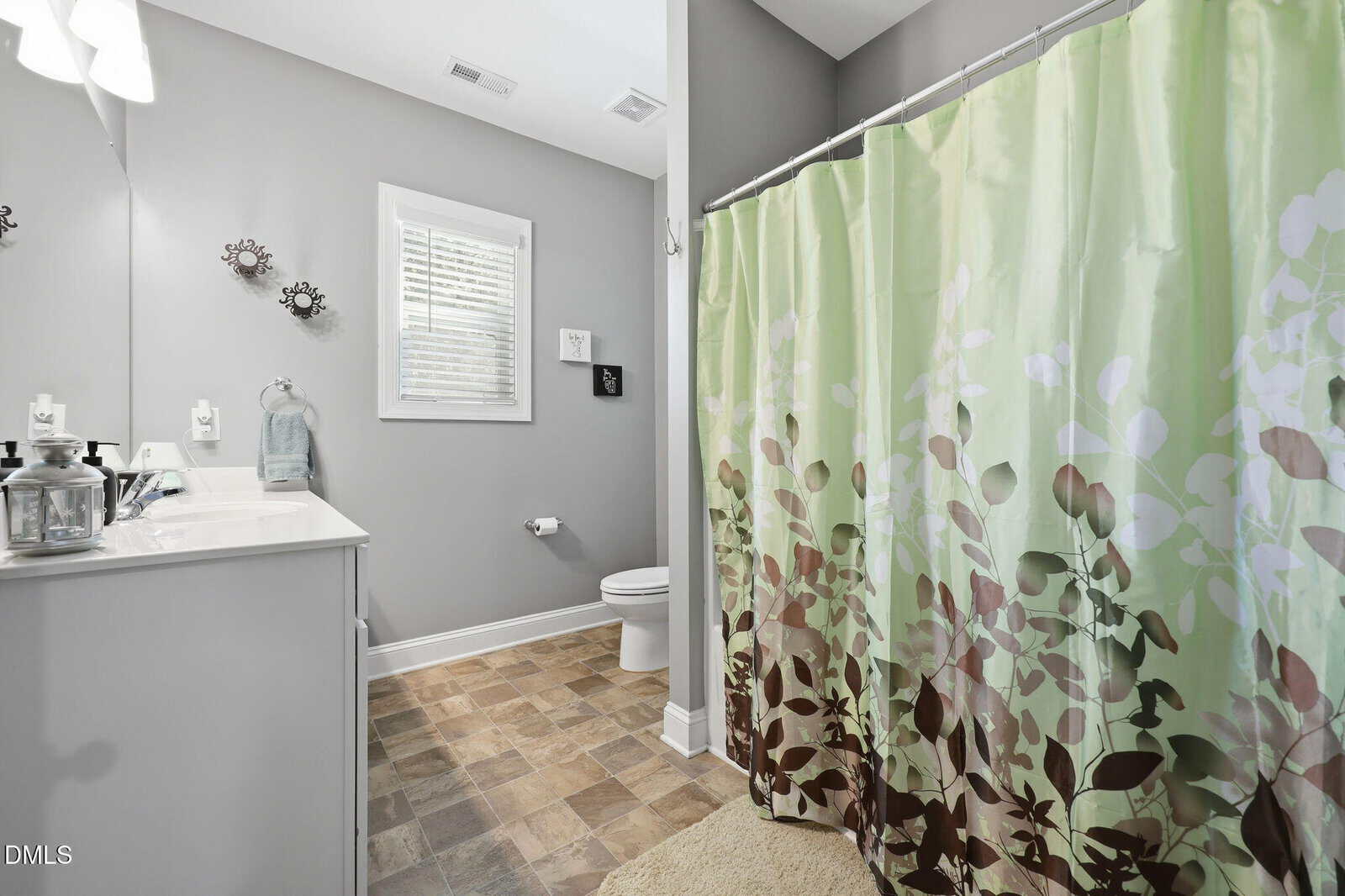 A modern bathroom with gray walls, a white vanity, a toilet, and a shower with a green and brown leafy-patterned curtain. There's a window above the toilet and decorative wall art near the mirror.