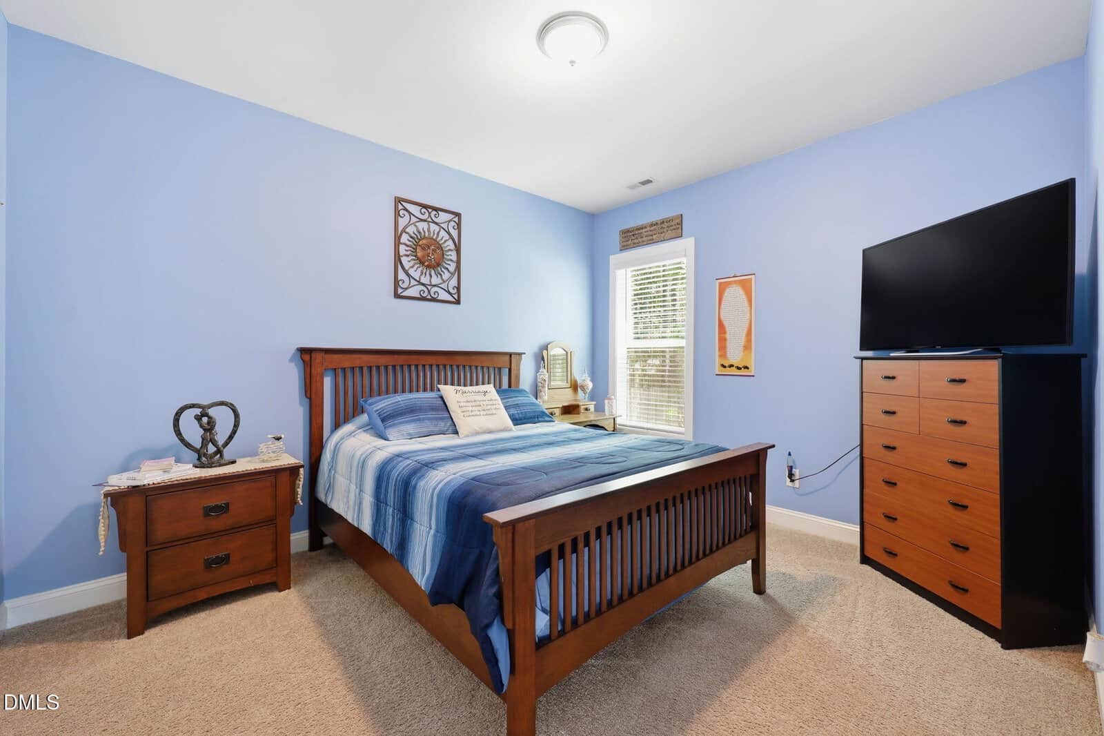 Bedroom with blue walls, a wooden bed with a blue plaid comforter, a nightstand, dresser with TV, carpeted floor, two windows, and framed wall art above the bed and between the windows.