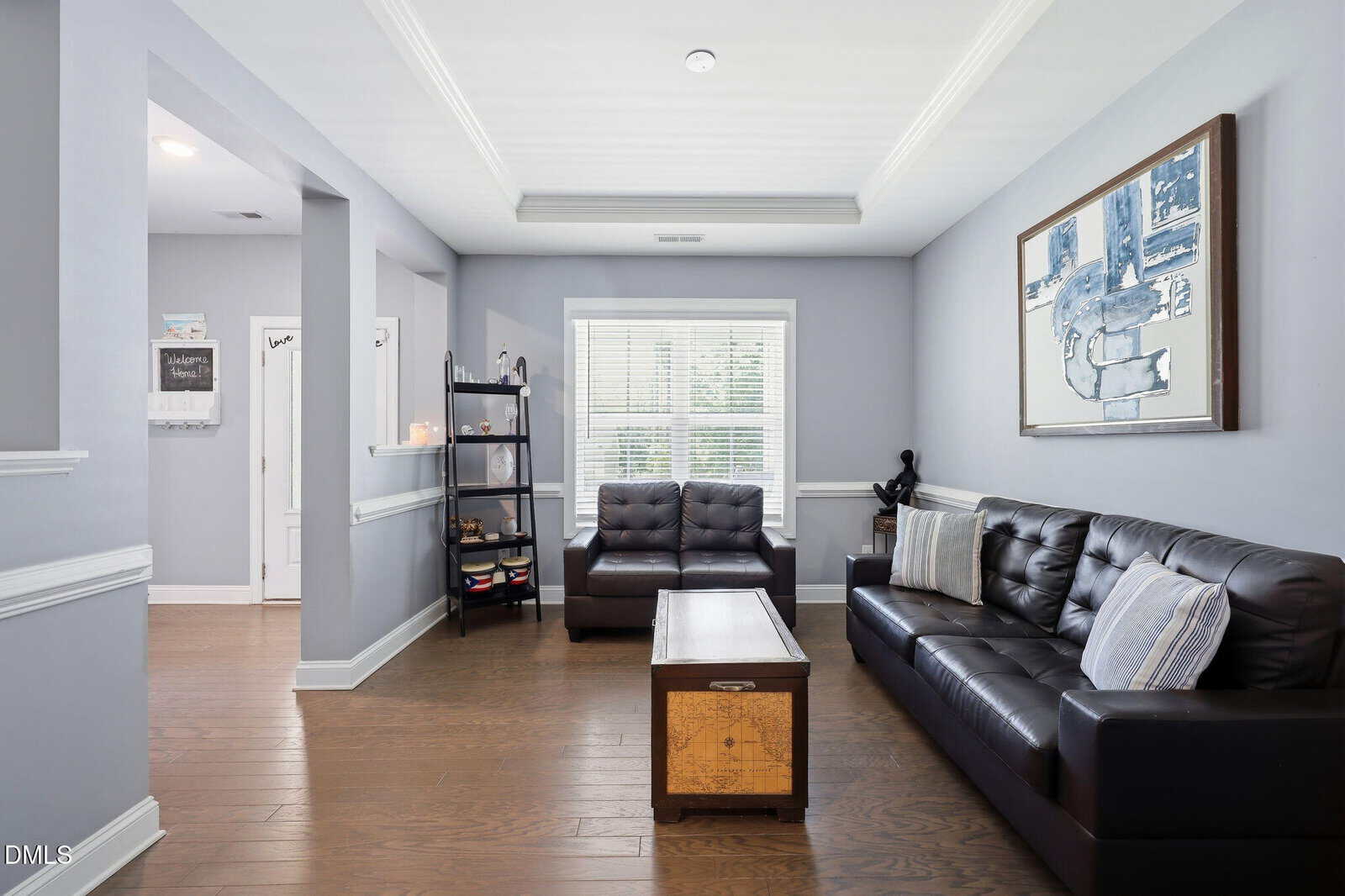 A modern living room with gray walls, dark leather sofas, a wooden coffee table, a bookshelf, and a large window with blinds letting in natural light.