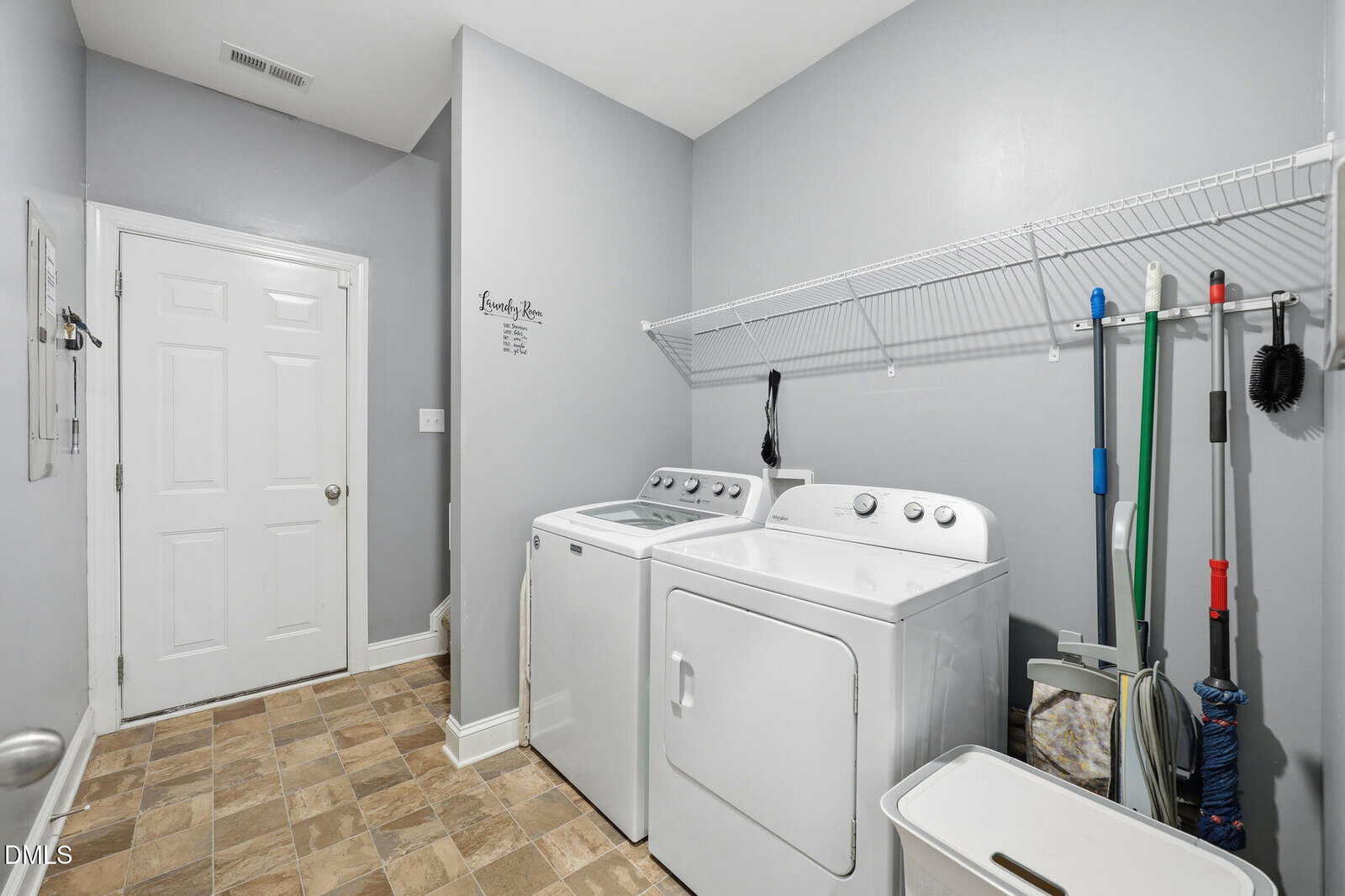 A laundry room with a washer and dryer, wire shelf above, cleaning tools, small utility bin, tile floor, and a white door on the left wall. Walls are painted light gray.