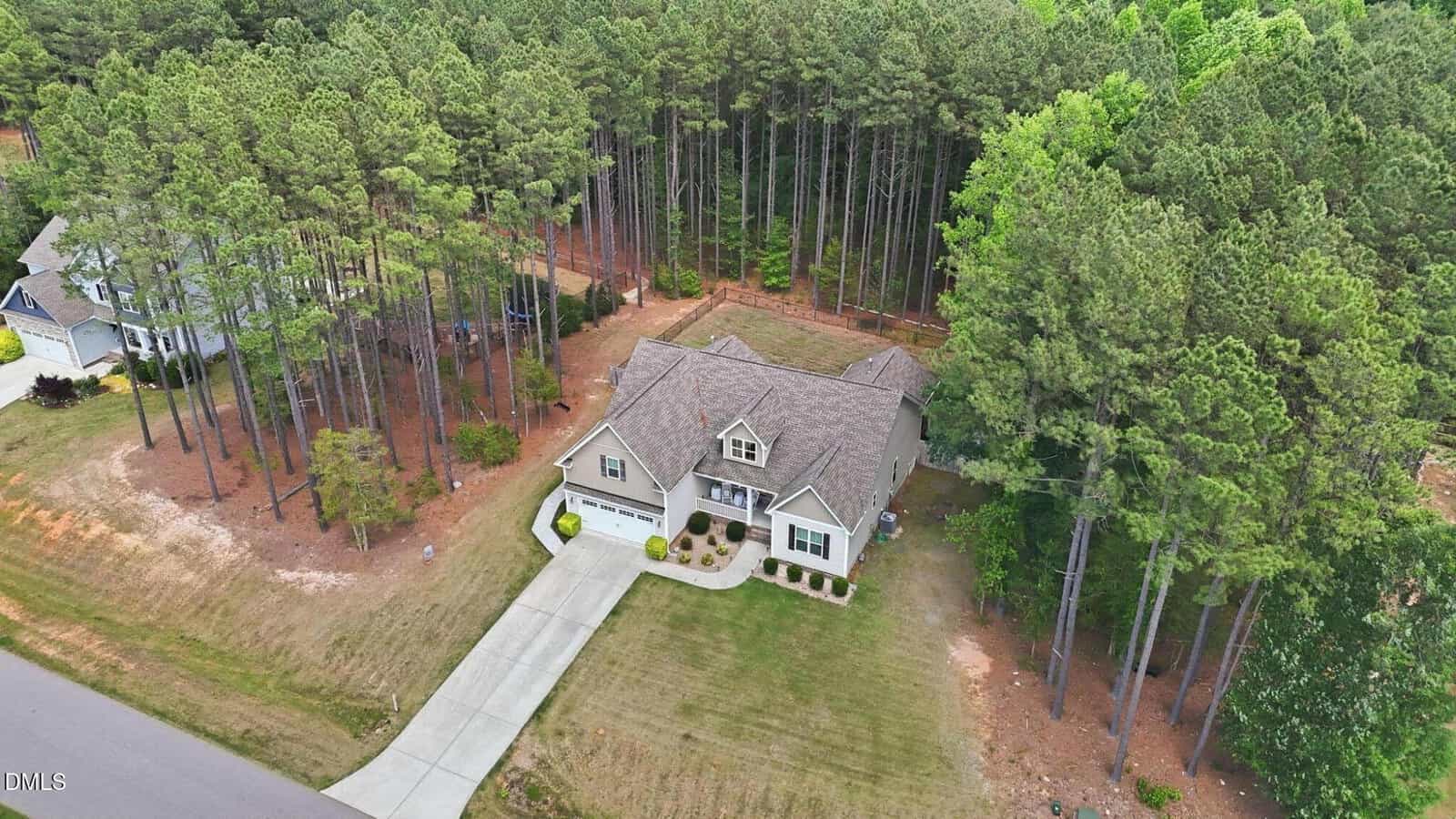 A two-story house with a two-car garage, beige exterior, and shingle roof sits beside a tall pine forest at 906 Weatherby Lane in Creedmoor. The lawn is patchy, and the home is bordered by a curved driveway leading to the street. Neighboring houses are visible.
