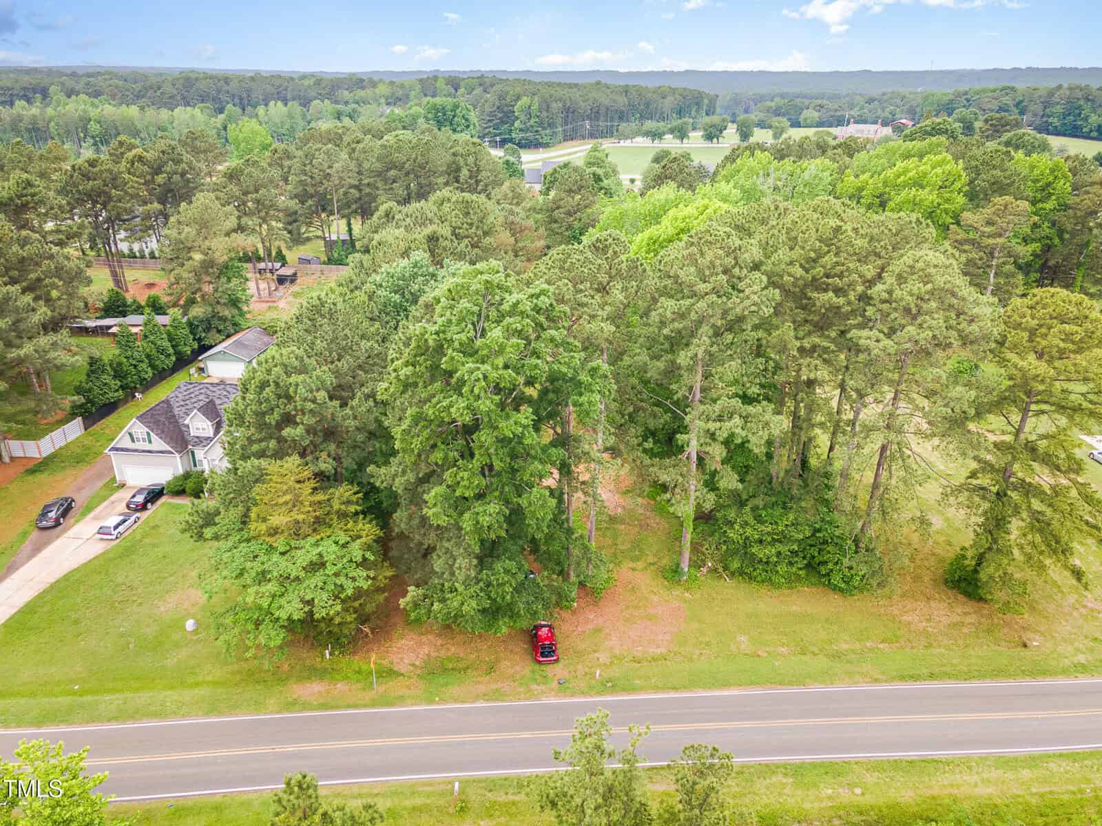 Aerial view of a wooded lot with tall trees next to a paved road at 63 Mount Olivet Church Road, Franklinton. A red car is parked under the trees, and a white house with a driveway is visible to the left, with more trees and a park-like area in the background.