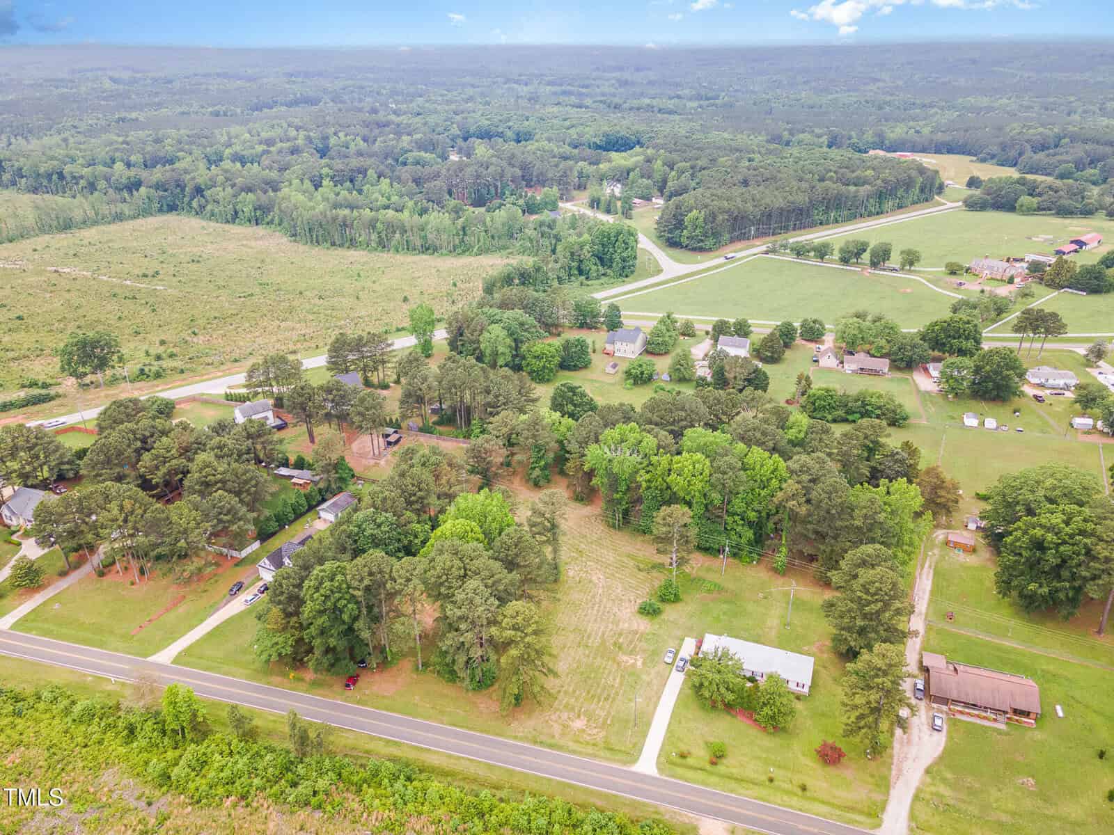 Aerial view of a rural area with scattered houses, roads, and green trees surrounded by open fields and forests under a partly cloudy sky.