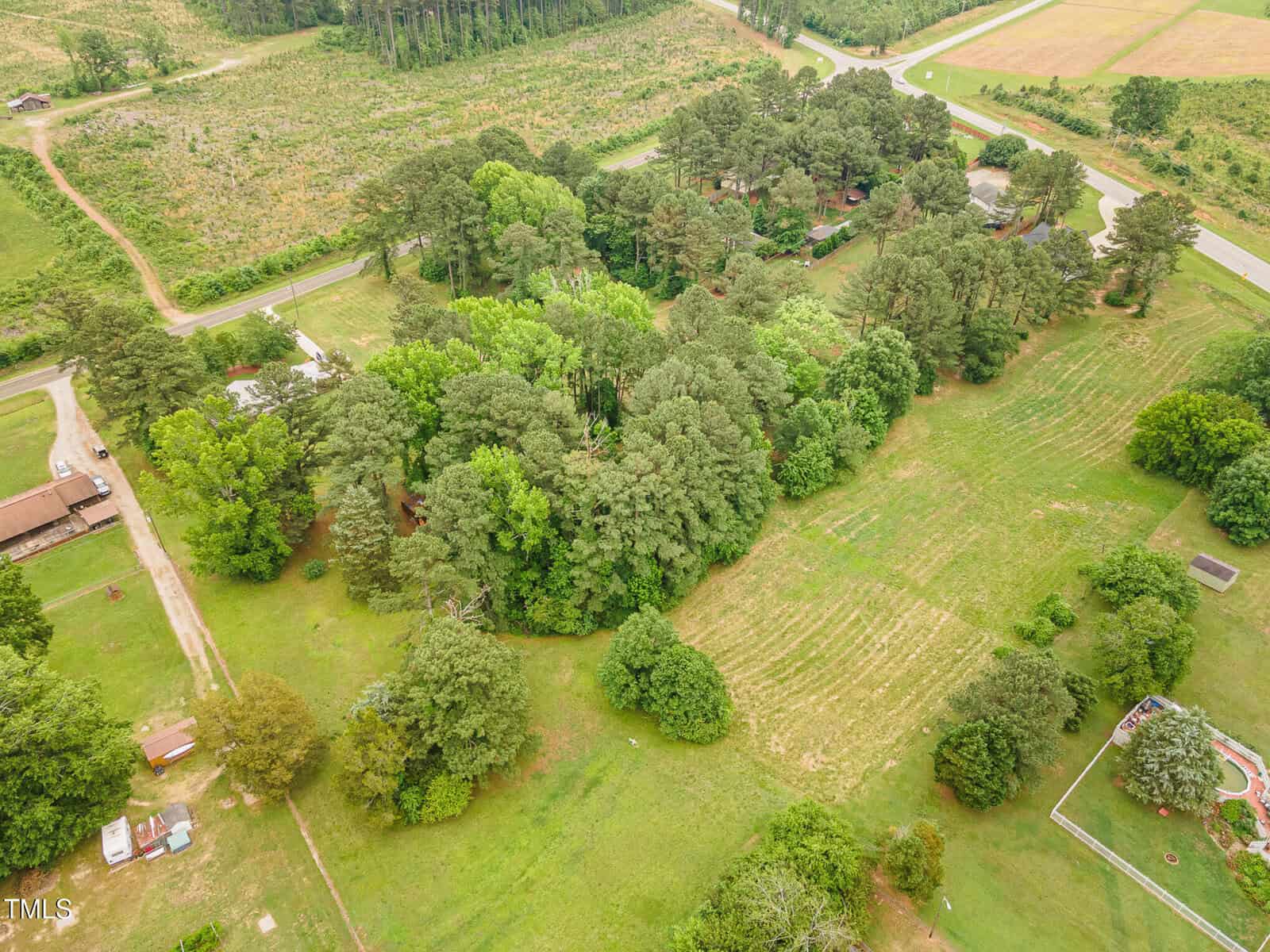 Aerial view of a rural landscape featuring clusters of trees, open grassy fields, a few houses, a garden area, and intersecting roads, surrounded by farmland and greenery.
