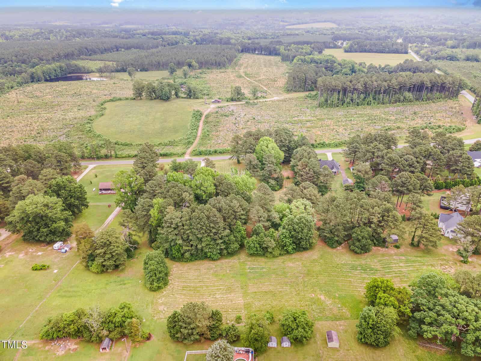 Aerial view of a rural landscape with fields, scattered trees, and several houses along a road surrounded by greenery and forested areas under a cloudy sky.