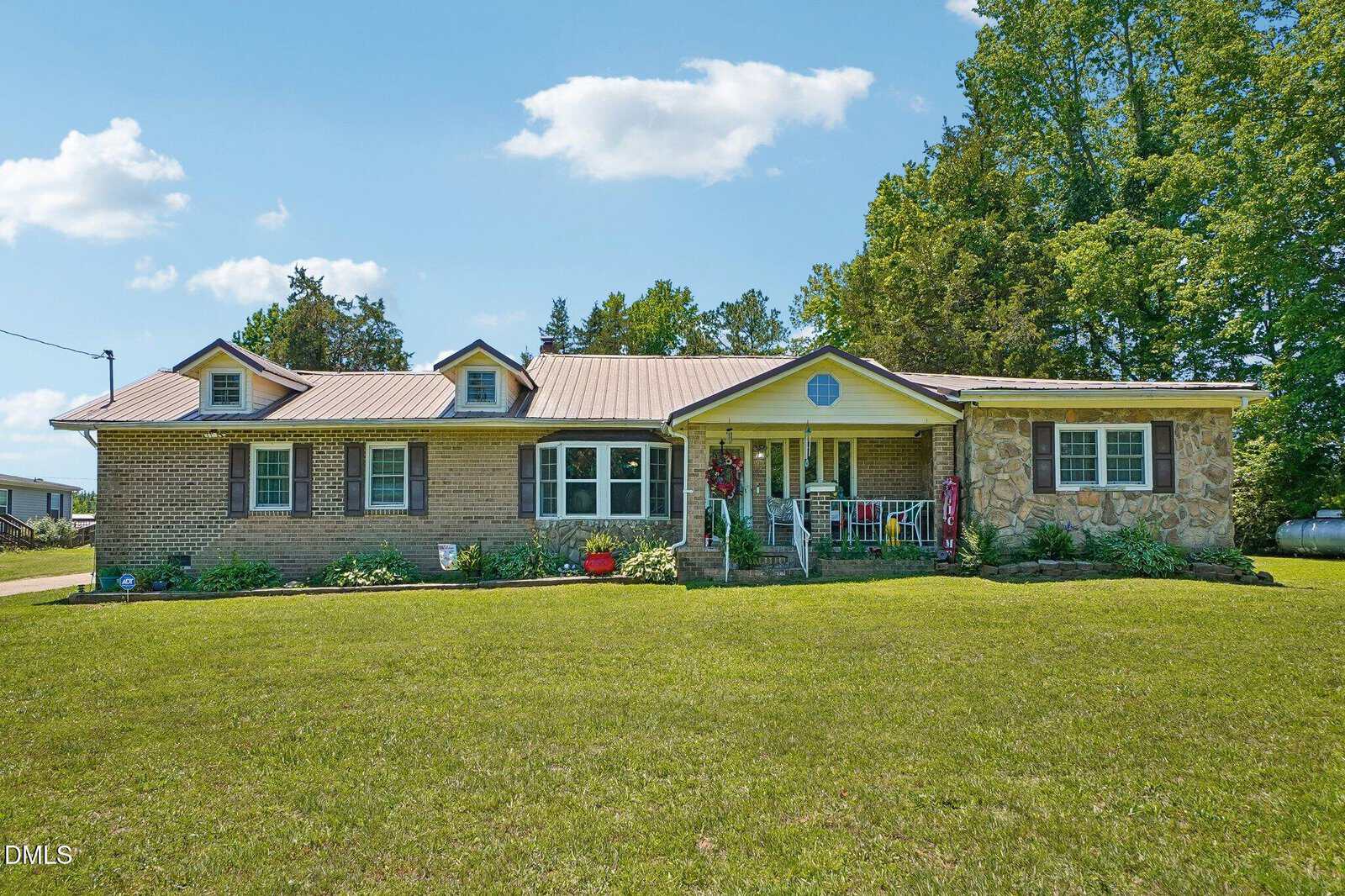 Single-story brick and stone house with three dormer windows, a covered front porch, green lawn, trees, and a bright, sunny sky with a few clouds.