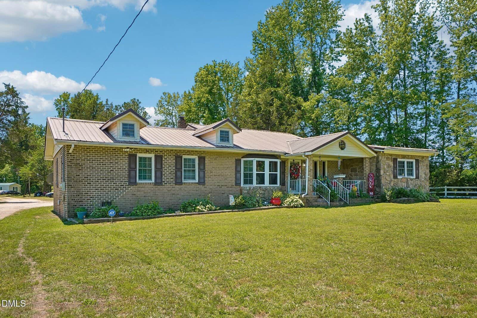 A single-story brick house with a metal roof, front porch, and dormer windows, surrounded by green grass and tall trees under a blue sky with scattered clouds.