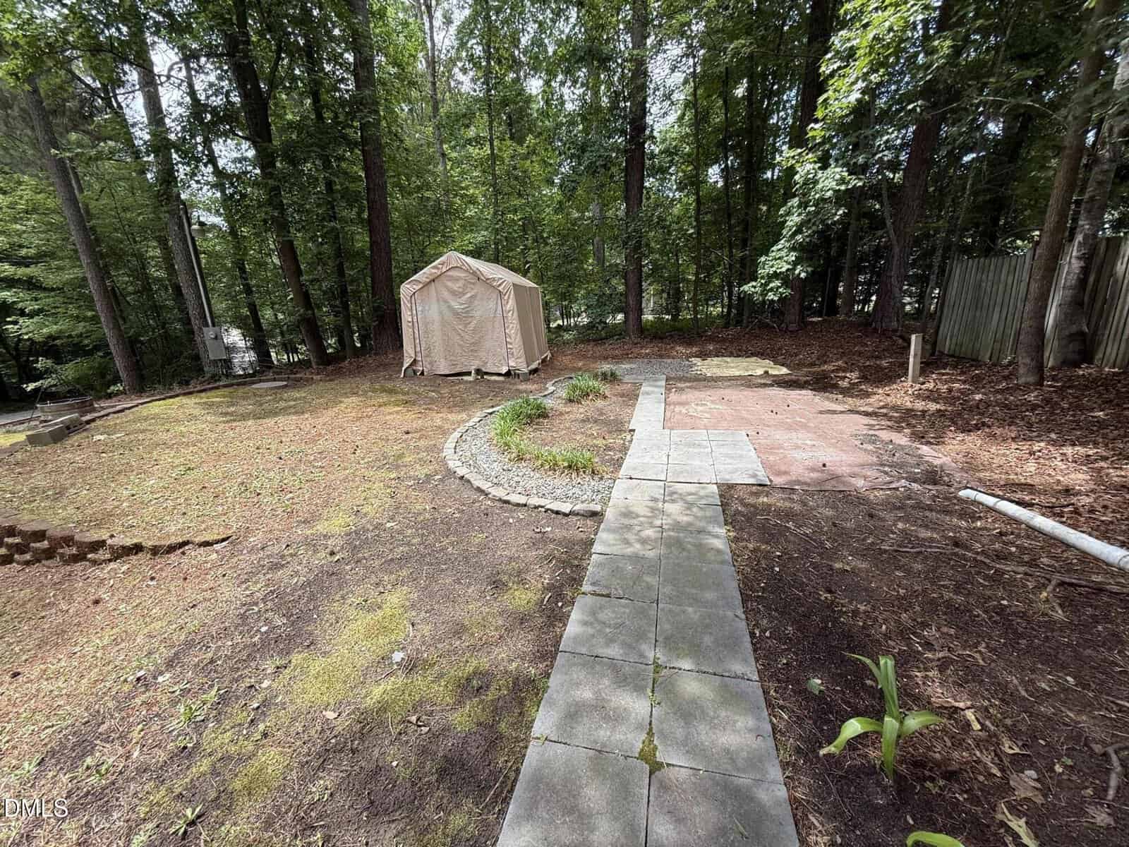 A backyard with a stone walkway leading to a beige tent under tall trees. There is a garden bed surrounded by rocks, patches of bare ground, and a wooden fence on the right.