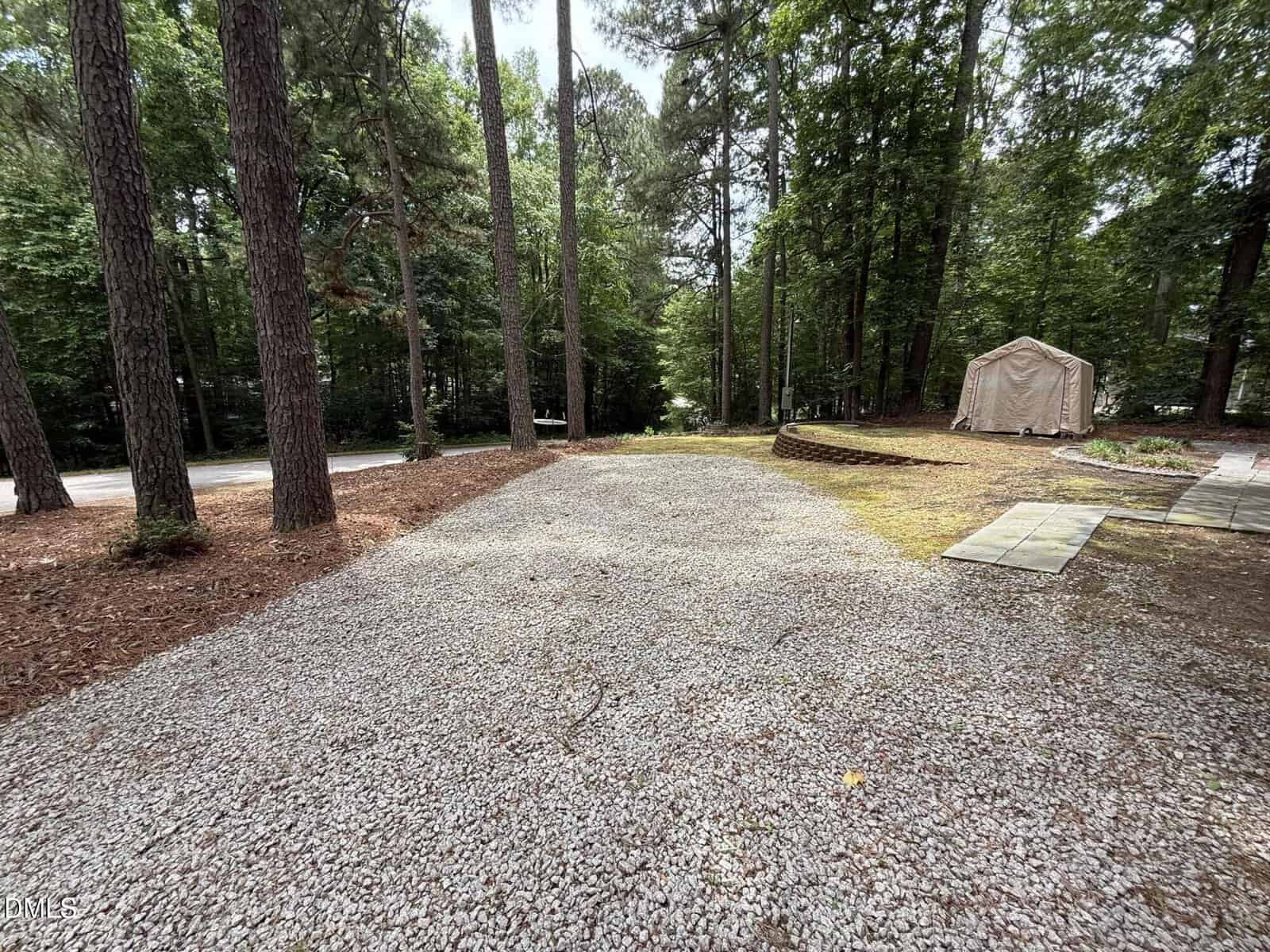 A gravel driveway surrounded by tall trees leads to a road. There is a beige tarp-covered structure on the right, bordered by a small retaining wall and stepping stones. Lush green forest fills the background.