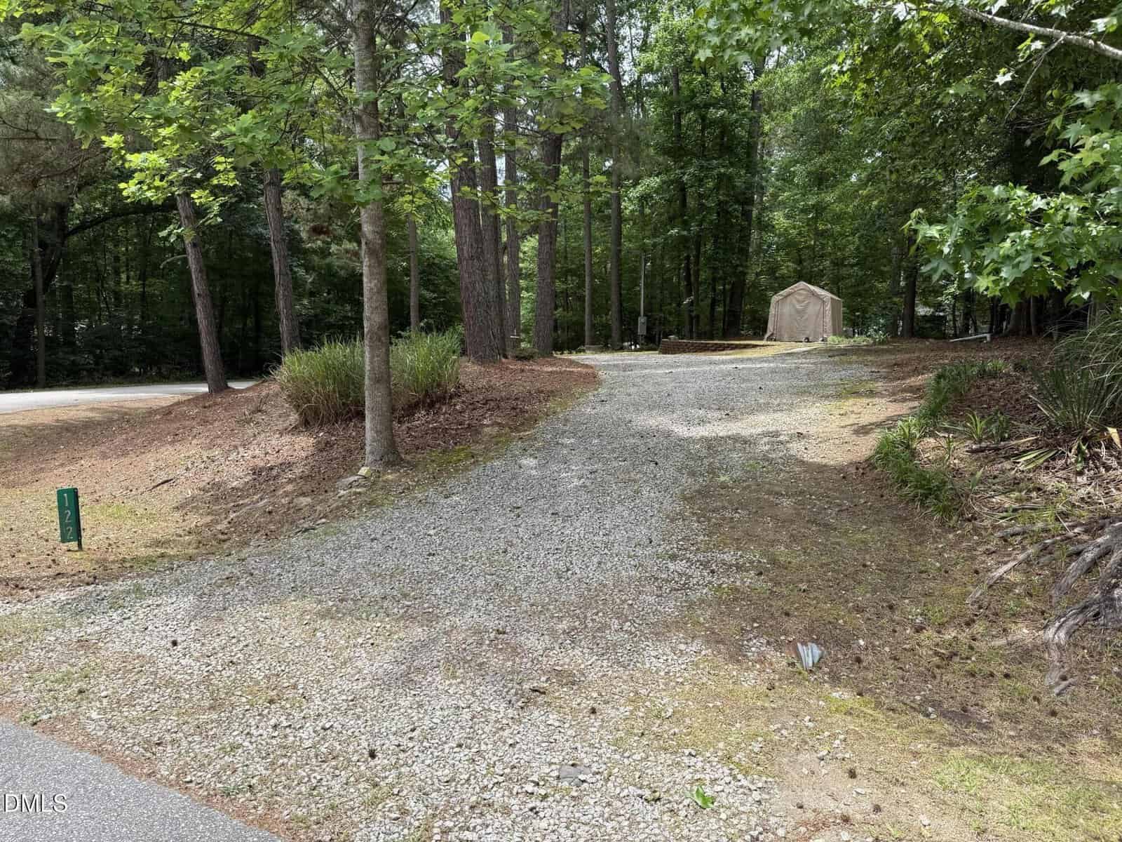 A gravel driveway at 122 Desoto Drive leads uphill through tall trees to a small beige shed in a wooded area in Louisburg, with grass and plants lining the sides and a paved road visible on the left.