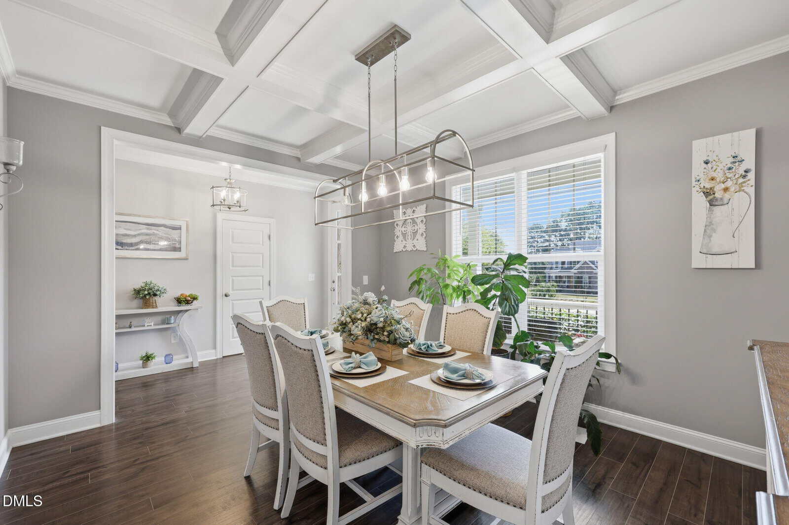 Bright dining room with a coffered ceiling, elegant chandelier, six upholstered chairs around a set dining table, large window letting in sunlight, potted plants, gray walls, hardwood floors, and decorative art.