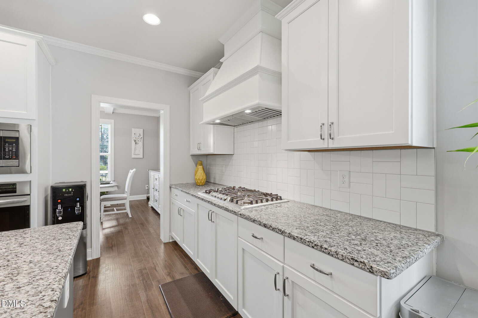 Modern kitchen with white cabinets, granite countertops, a gas cooktop, stainless steel appliances, and wood flooring; a dining area is visible through a doorway in the background.