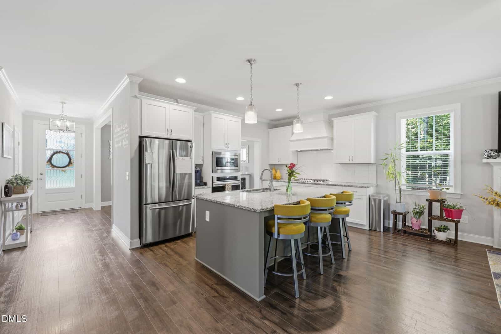 Modern kitchen with white cabinets, stainless steel appliances, a large island with a granite countertop, three yellow barstools, pendant lights, and wood flooring. Bright, airy space with plants and windows.