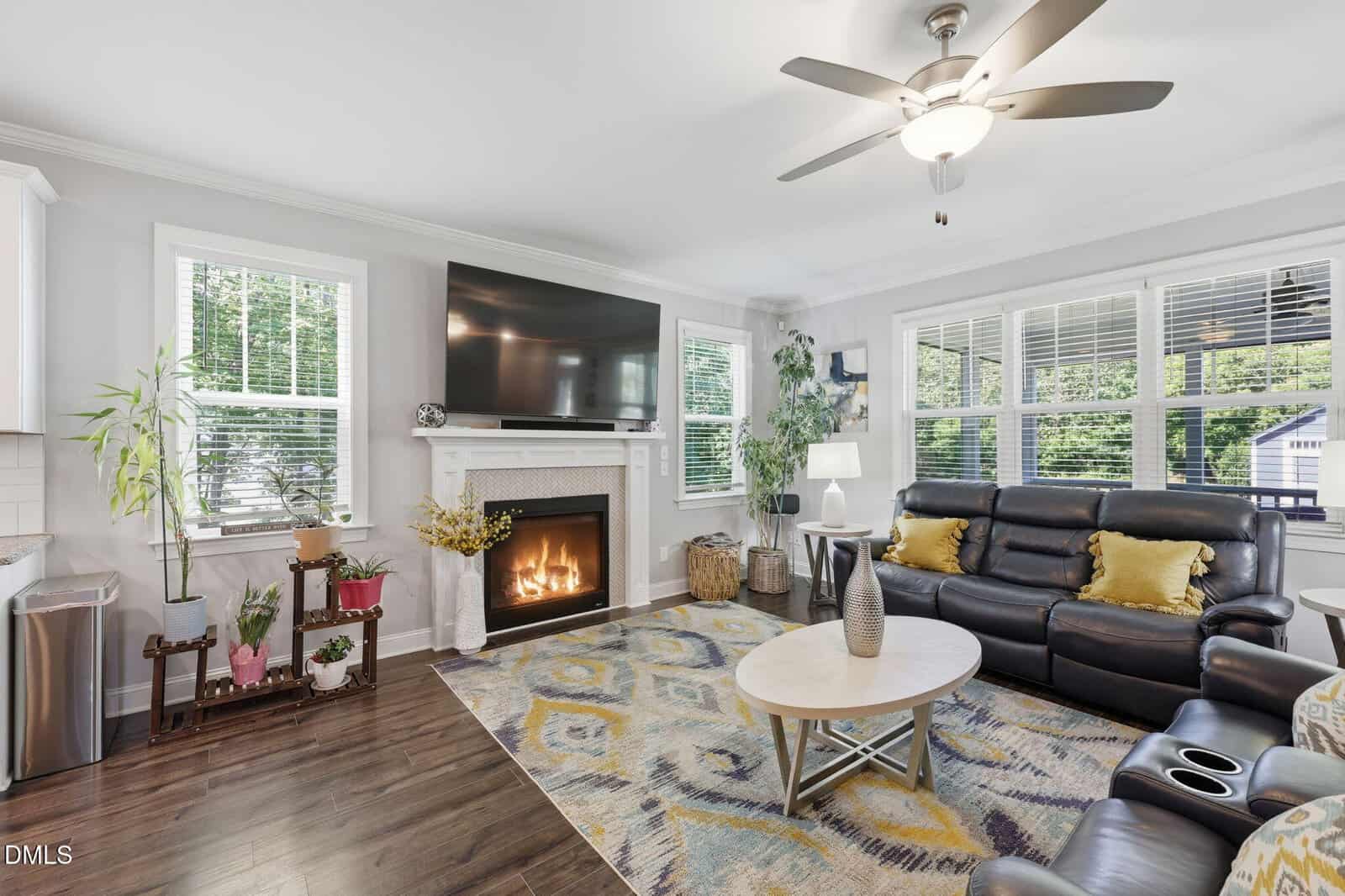 Modern living room with a black leather sofa, yellow pillows, a round coffee table, fireplace with fire, large wall-mounted TV, plants, and large windows letting in natural light. Ceiling fan and hardwood floors are visible.