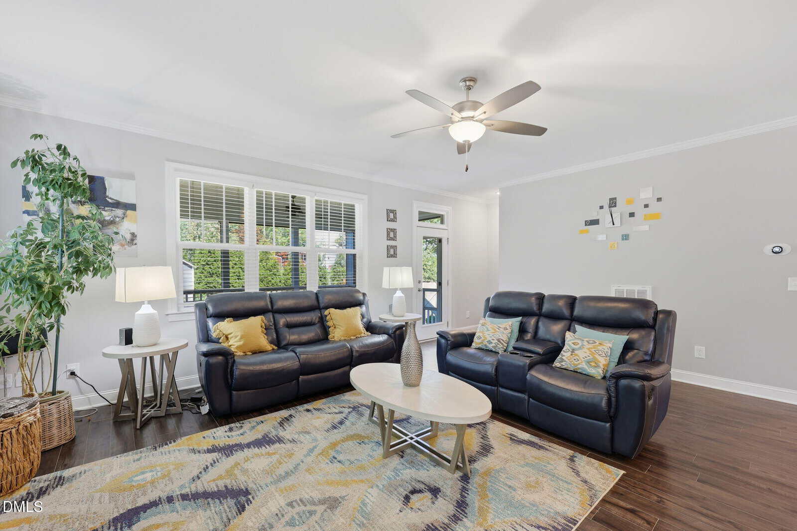 Bright living room with two black leather sofas, a patterned rug, a round coffee table, large windows, indoor plants, and wall art. Ceiling fan with light and hardwood flooring complete the modern, cozy space.