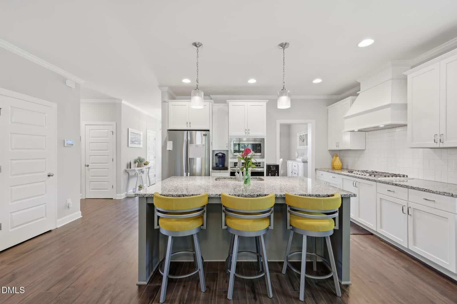 Modern kitchen with white cabinets, stainless steel appliances, a large island with a granite countertop, yellow barstools, pendant lights, and wood flooring. A vase with flowers sits on the island.