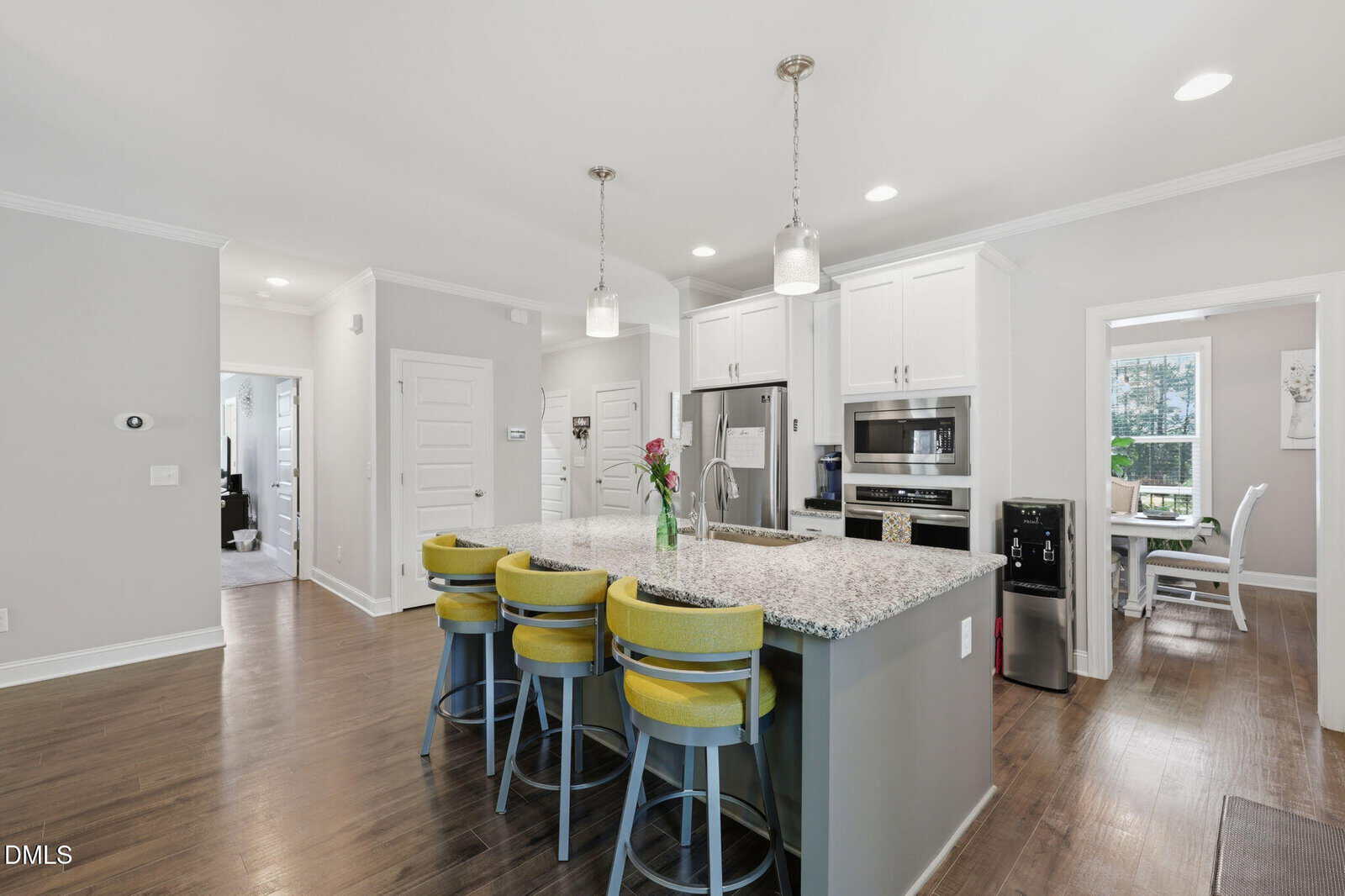 Modern kitchen with a large granite island, four yellow barstools, stainless steel appliances, white cabinets, pendant lights, and wooden floors. A vase with flowers sits on the island. Dining area visible in the background.