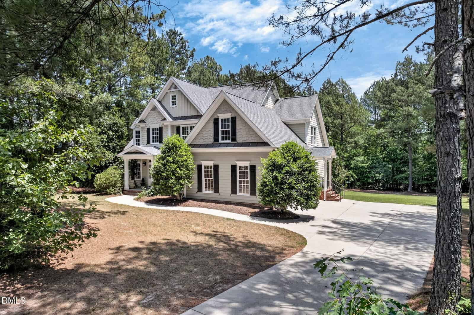 A large two-story house with gray siding, white trim, and a covered front porch sits among tall trees. A curved driveway leads to the entrance, and manicured shrubs are planted near the front of the house.