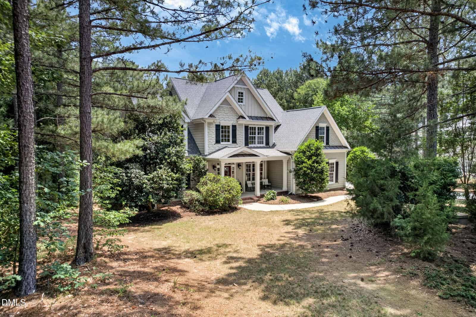 A two-story suburban house with light gray siding, white trim, and a gabled roof sits surrounded by tall pine trees and shrubs under a blue sky with scattered clouds.