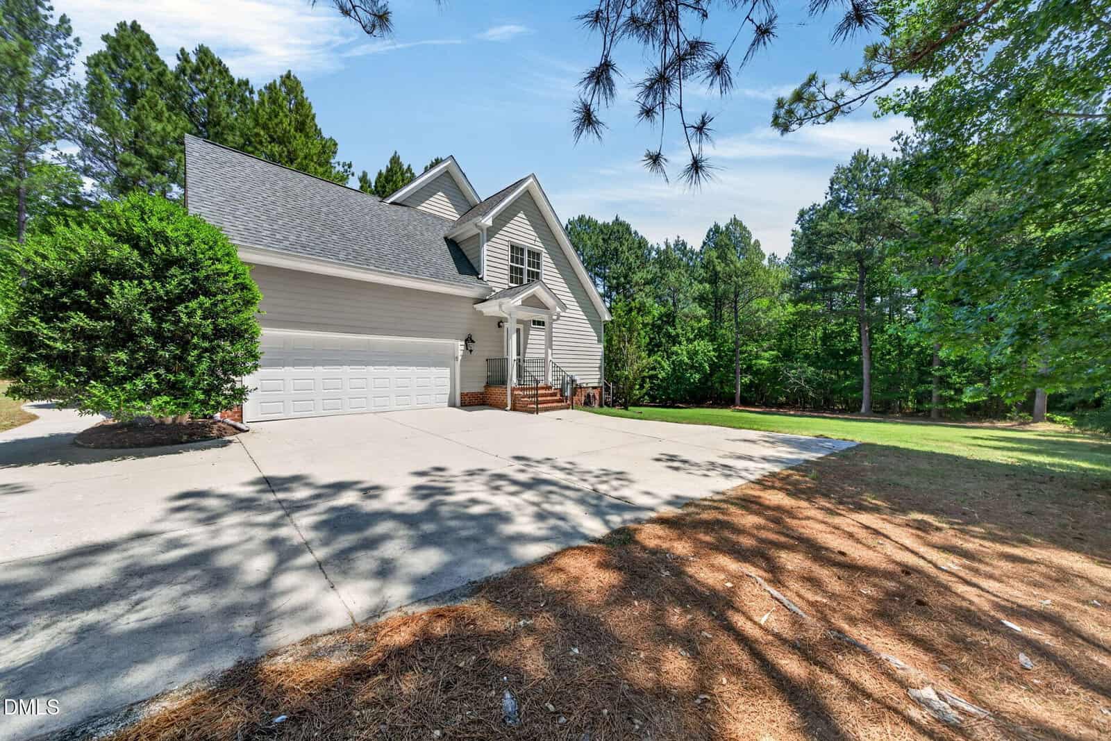A white two-story house with a large driveway, attached two-car garage, brick steps at the front entrance, surrounded by pine trees and a green lawn under a partly cloudy sky.
