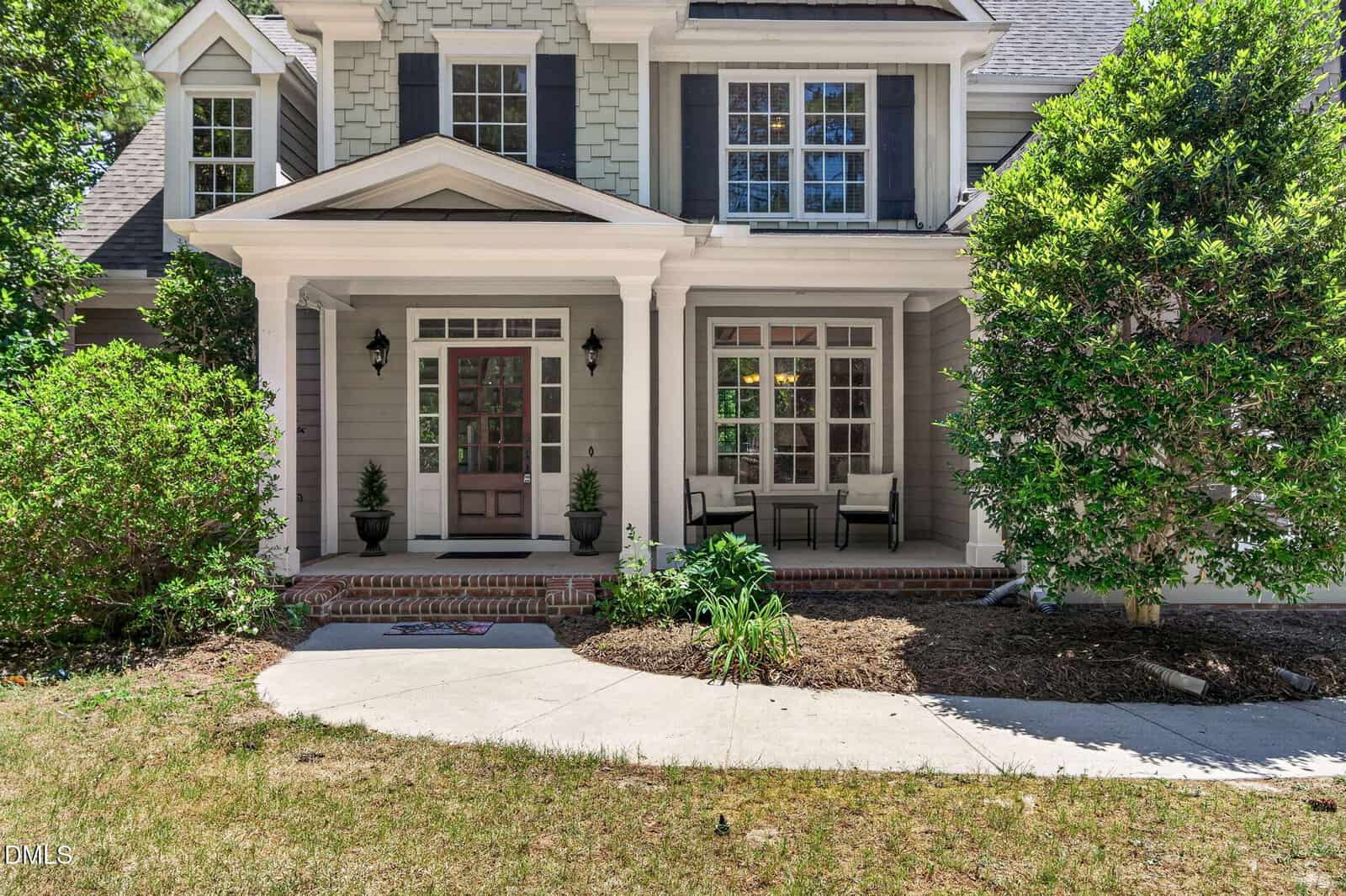 A two-story house with light gray siding, white trim, black shutters, and a covered front porch featuring potted plants, a bench, and two chairs. A curved walkway leads to the entrance, surrounded by green shrubs and trees.