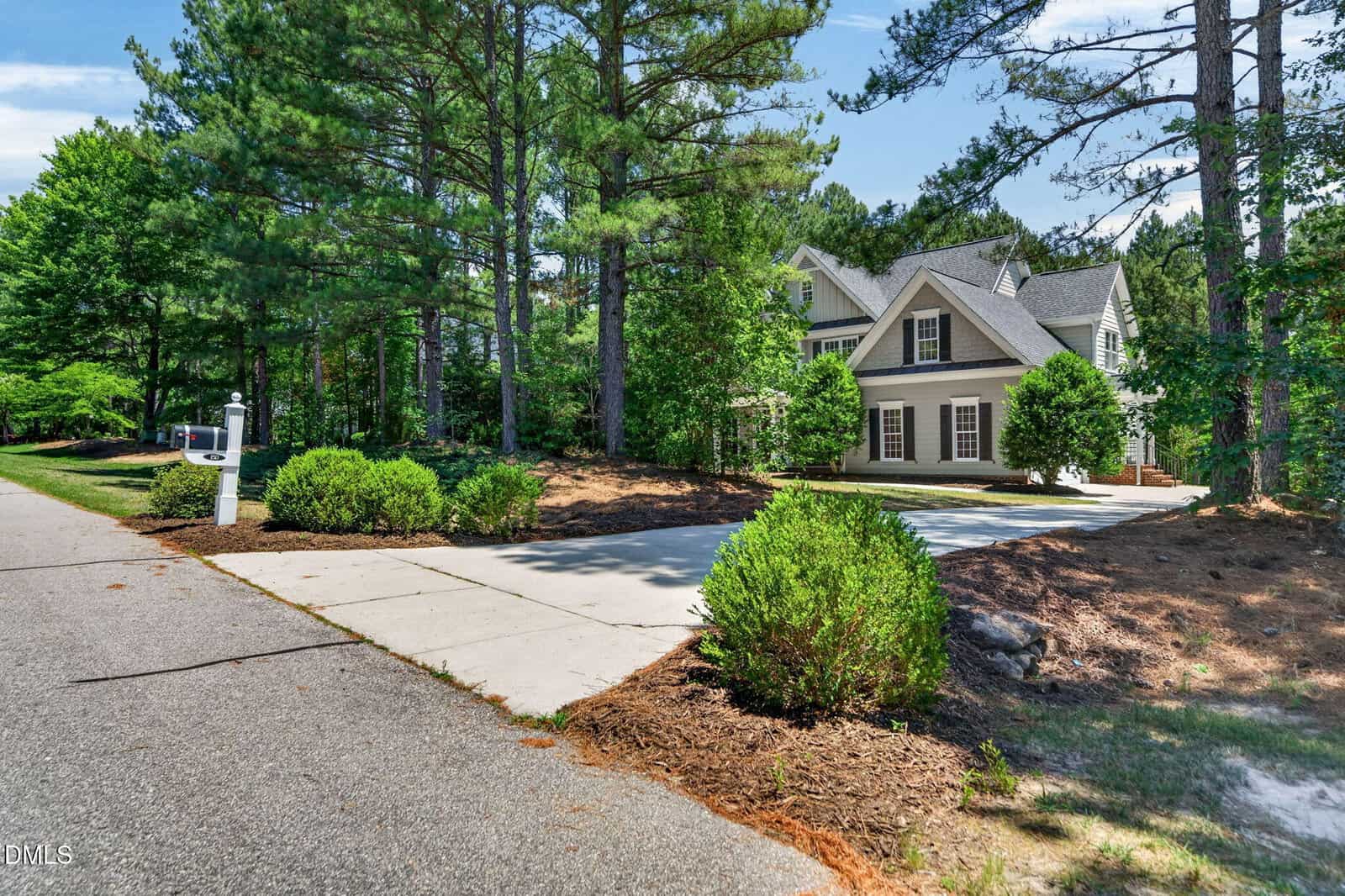 A two-story house with beige siding and white trim sits surrounded by tall pine trees. The home has manicured bushes, a wide driveway, and is located next to a quiet, tree-lined street.