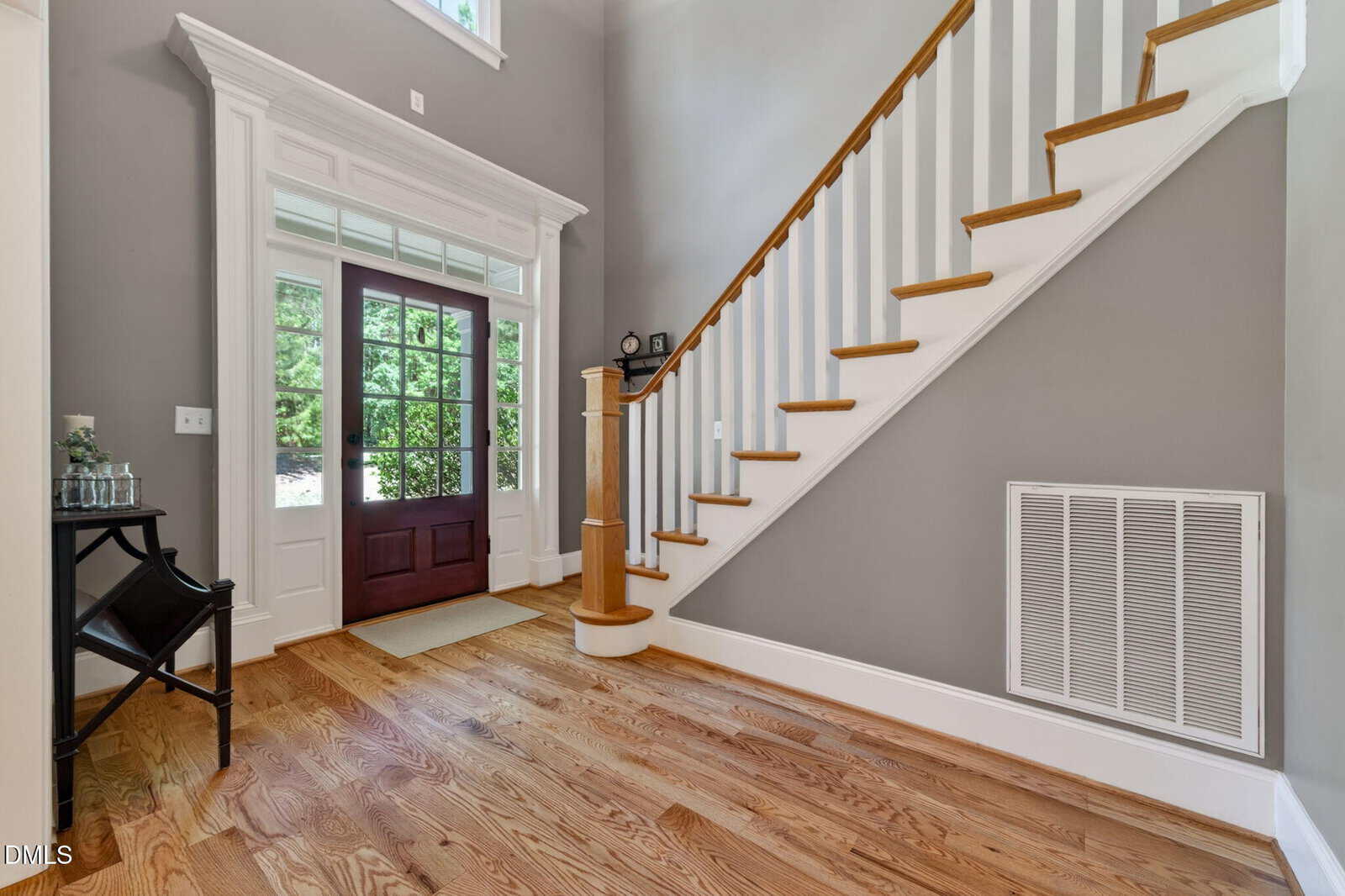 A bright foyer with wood floors, a staircase with white spindles and a wooden handrail, and a glass front door. A small table with glassware is to the left, and there is a large vent on the right wall.
